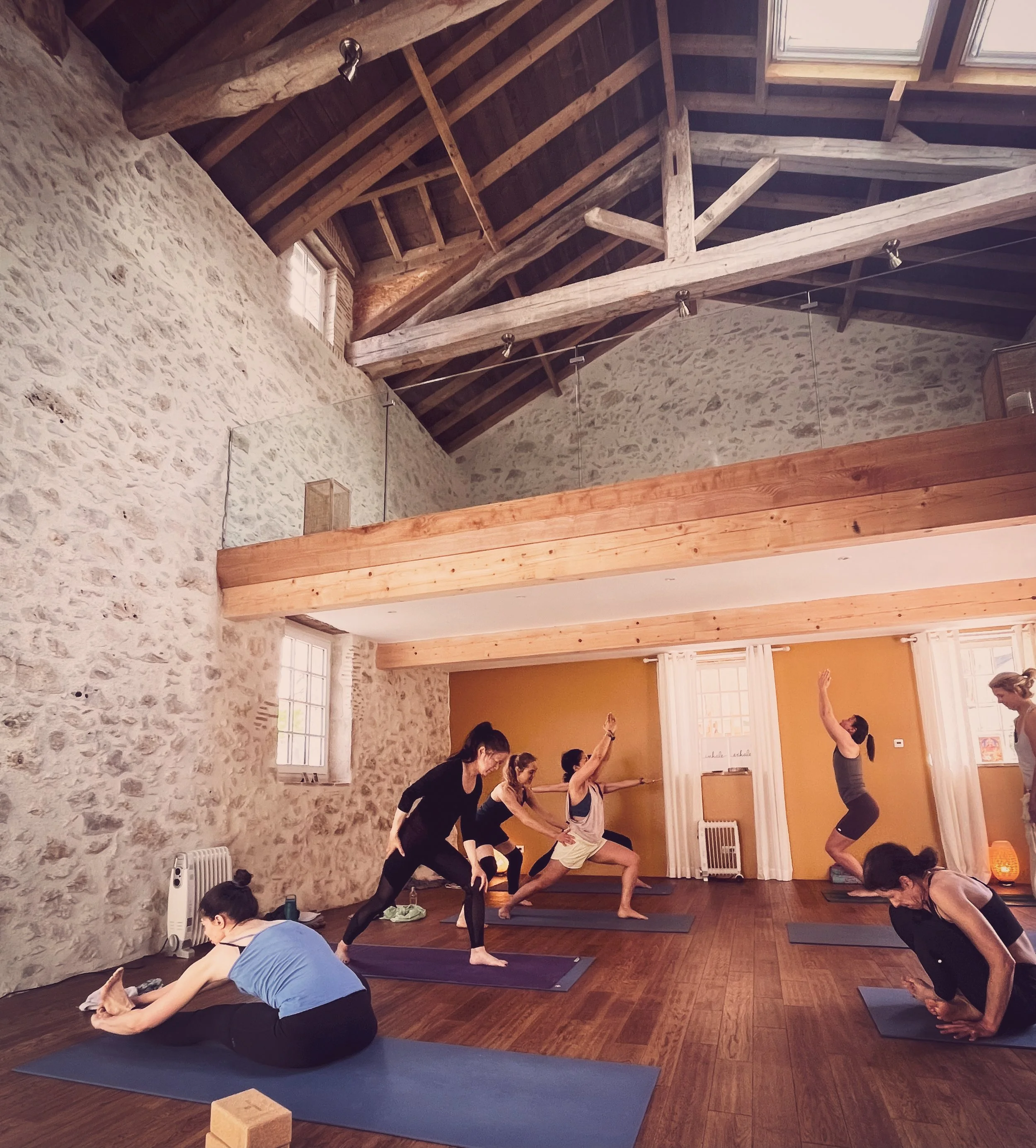 Groupe de personnes pratiquant du yoga dans une salle avec murs en pierre, poutres en bois au plafond et fenêtres naturelles.