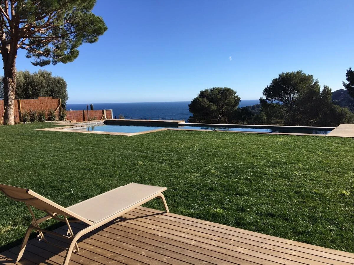 Une terrasse en bois avec un transat, un jardin bien entretenu, une piscine et une vue sur la mer avec des arbres et un ciel bleu.