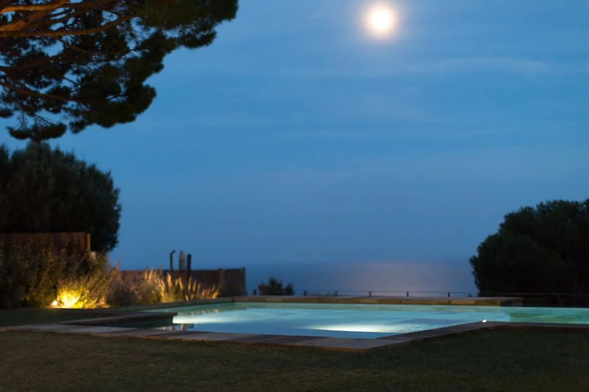 Piscine extérieure illuminée la nuit, avec la lune dans un ciel clair, entourée d'arbres et de haies.