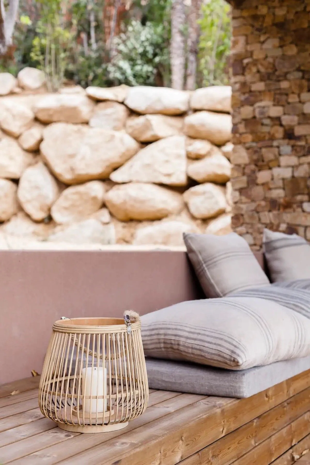 Coin salon extérieur avec un canapé beige à coussins rayés, une table en bois avec une lanterne en forme de cage contenant une bougie blanche, mur de pierre en fond et une banquette en pierre naturelle.