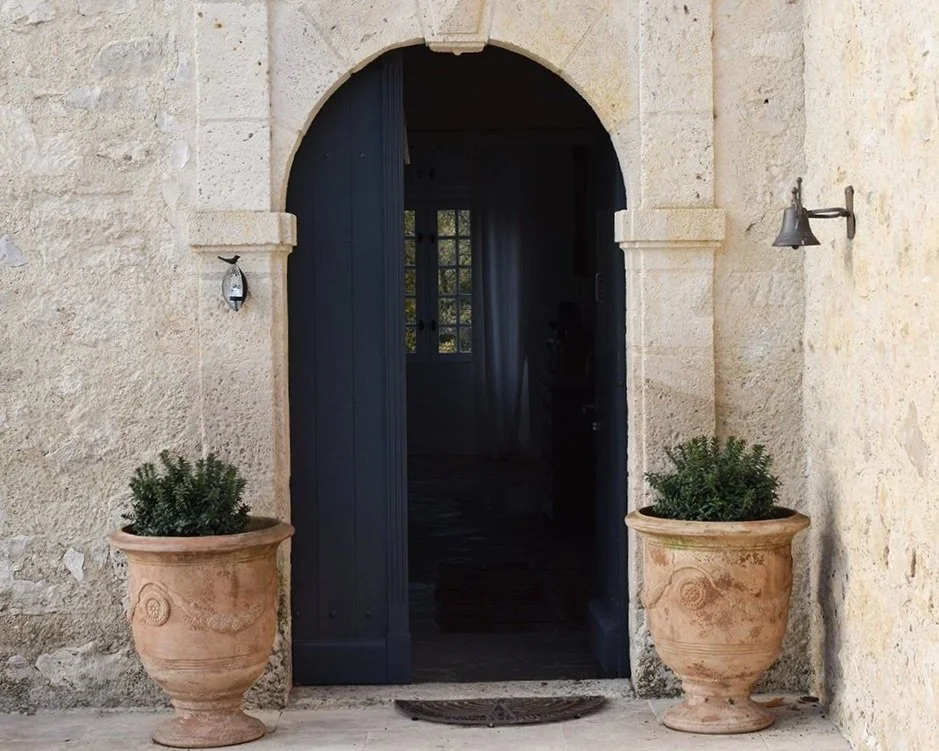 Porte en bois sombre encadrée d'une arche en pierre, flanquée de deux grands pots en terre cuite avec des arbustes. Deux appliques murales modernes de chaque côté de la porte, façade en pierre beige rustique.