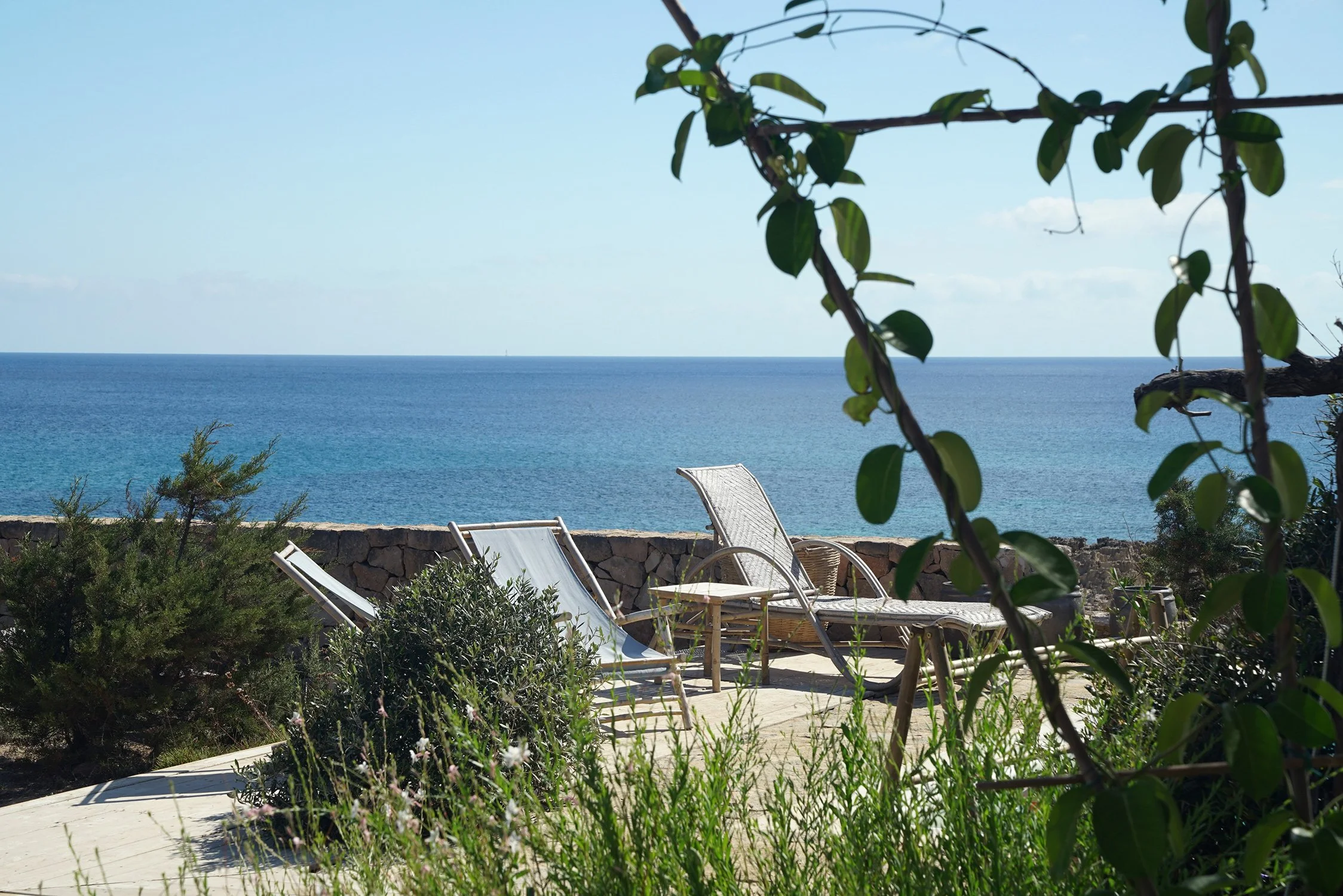 Chaises longues en bois regardant la mer dans un cadre naturel avec des plantes et un mur en pierre.