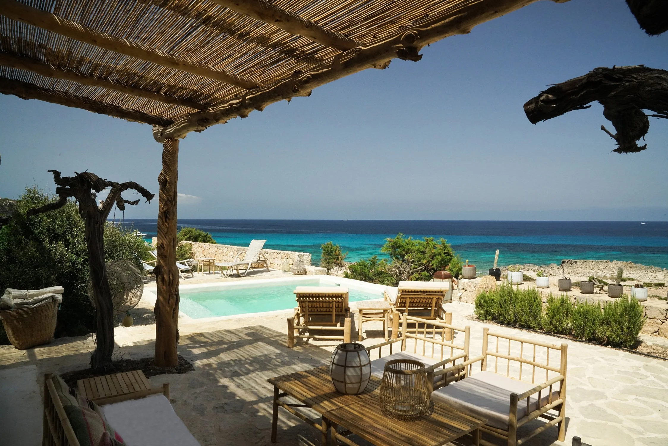 Terrasse avec mobilier en bois, vue sur une piscine, des plantes et la mer, sous un auvent en bois et paille.