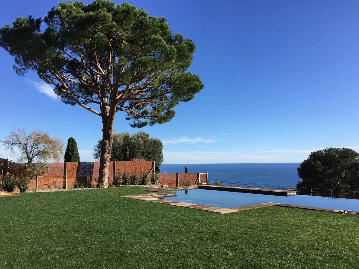 Jardin avec piscine, grand arbre vert, clôture en bois, vue sur la mer claire et ciel bleu.
