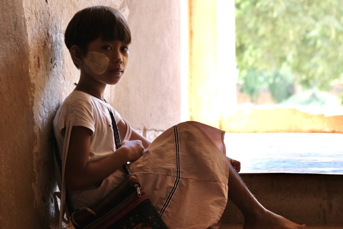 Une jeune fille assise à l'intérieur, près d'une fenêtre, portant une robe beige et un sac en bandoulière, avec un patch de pâte ou de pansement sur le visage.