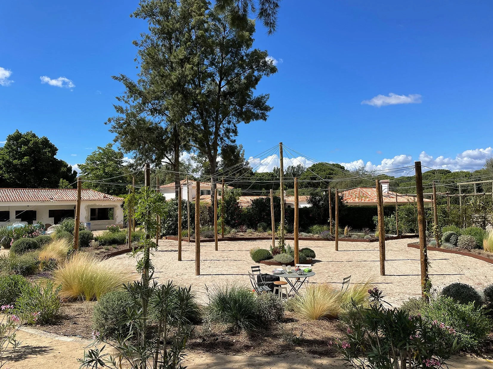 Jardin avec structure en bois pour le yoga ou la méditation, sur fond de maisons avec toits en tuiles, sous un ciel bleu avec quelques nuages.