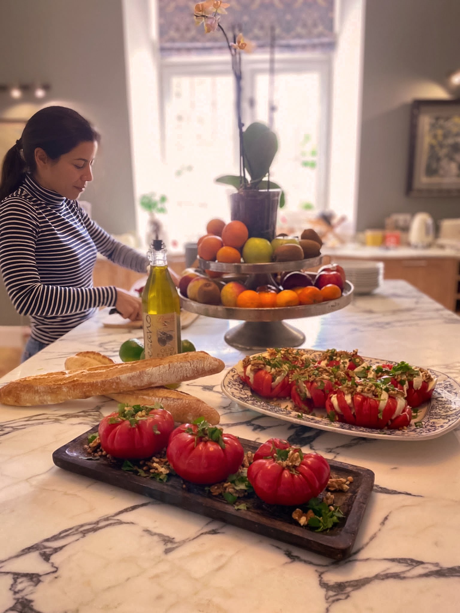 Une cuisine avec une femme qui prépare des aliments. Sur le plan de travail, il y a des tomates farcies, du pain baguette, une bouteille d'huile d'olive et un plateau de fruits avec des pommes et oranges. En arrière-plan, il y a une étagère avec un v