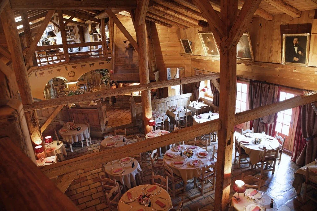 Salle à manger en bois dans un style rustique avec des tables préparées pour un repas, nappes blanches, et éclairage chaleureux, dans un bâtiment en bois avec des portraits accrochés aux murs.