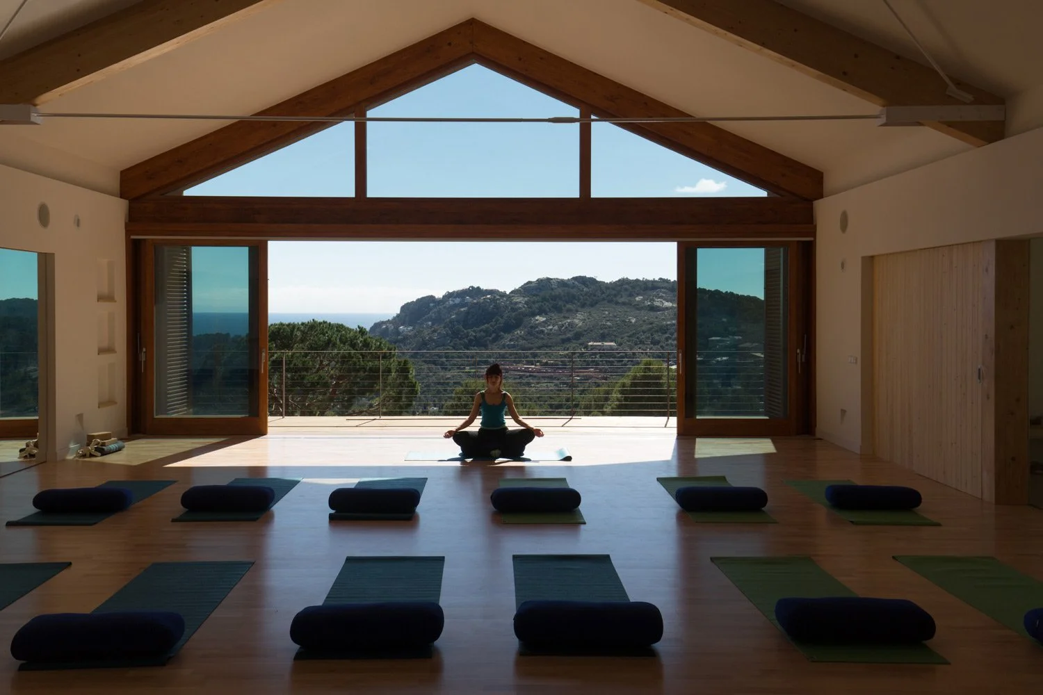 Une personne faisant de la méditation dans une salle de yoga avec vue sur la montagne et la mer, des tapis de yoga et des coussins