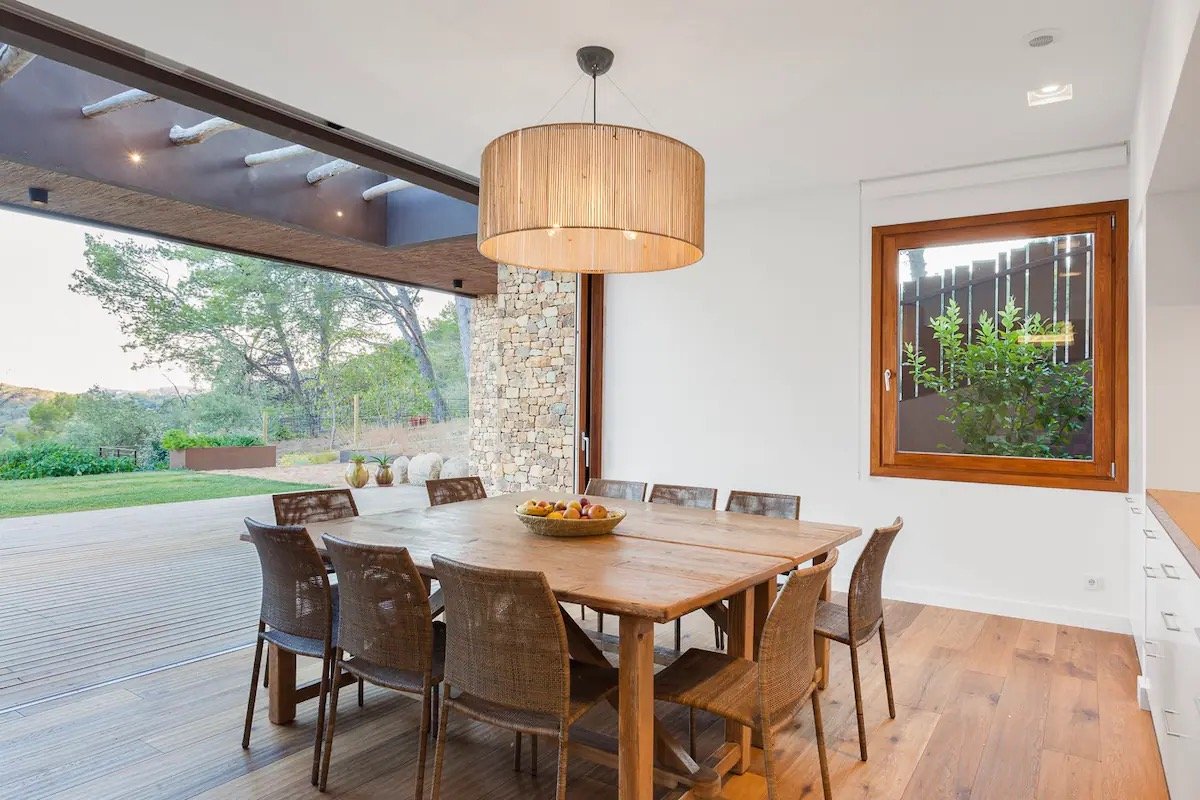 Salle à manger avec une table en bois, neuf chaises, un luminaire suspendu en forme de cylindre en rotin, une grande fenêtre donnant sur un espace extérieur avec des arbres.