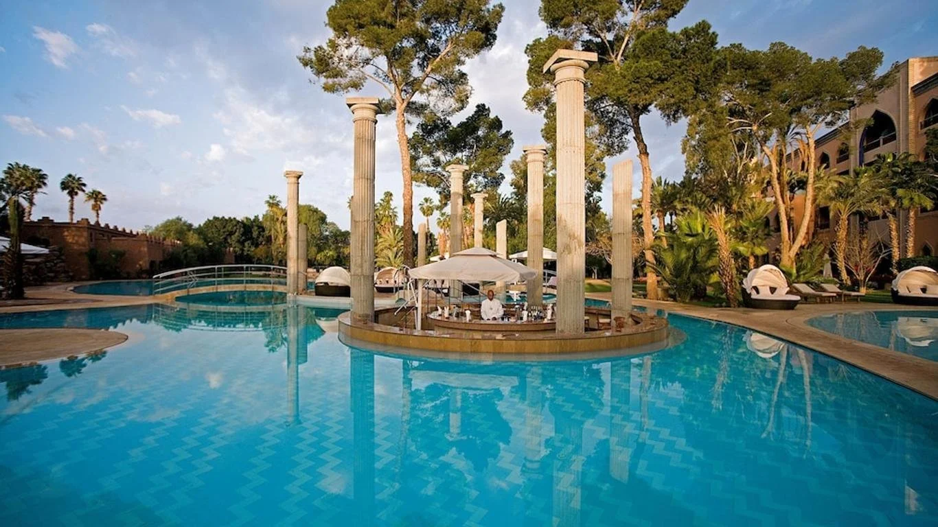 Une piscine à débordement entourée de colonnes antiques et de palmiers, avec un personnel de service sous un parasol blanc.