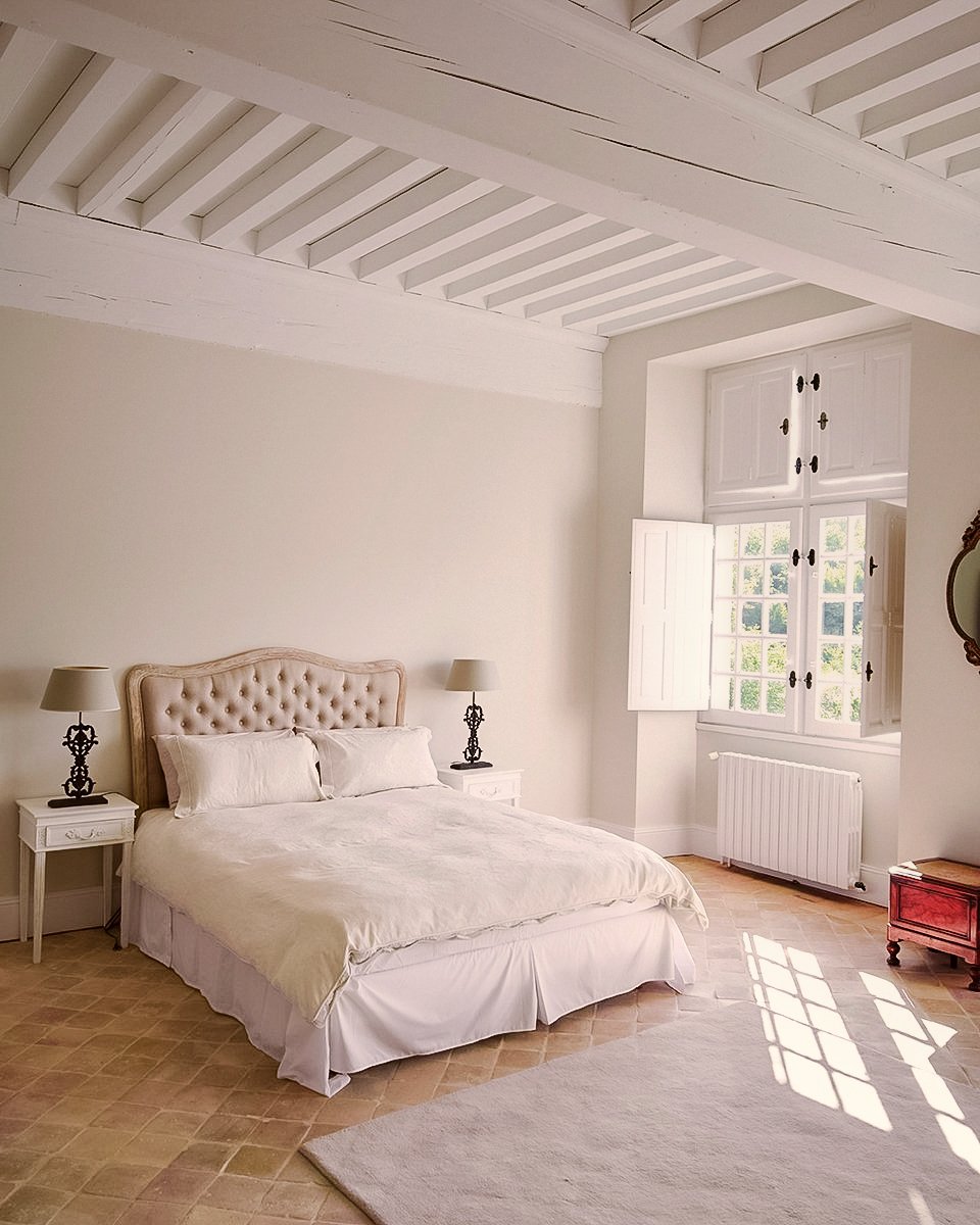 Chambre à coucher lumineuse avec un lit beige à tête capitonnée, deux lampes de chevet, une fenêtre avec volets blancs, un radiateur, un tapis beige et un petit meuble rouge.