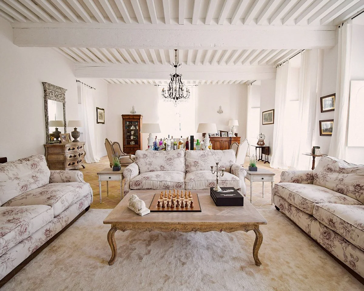 Salon lumineux avec trois canapés fleuris, une table avec un échiquier, un chandelier et un livre, et meuble ancien. Décoration élégante avec lampes, tableaux et chandelier.