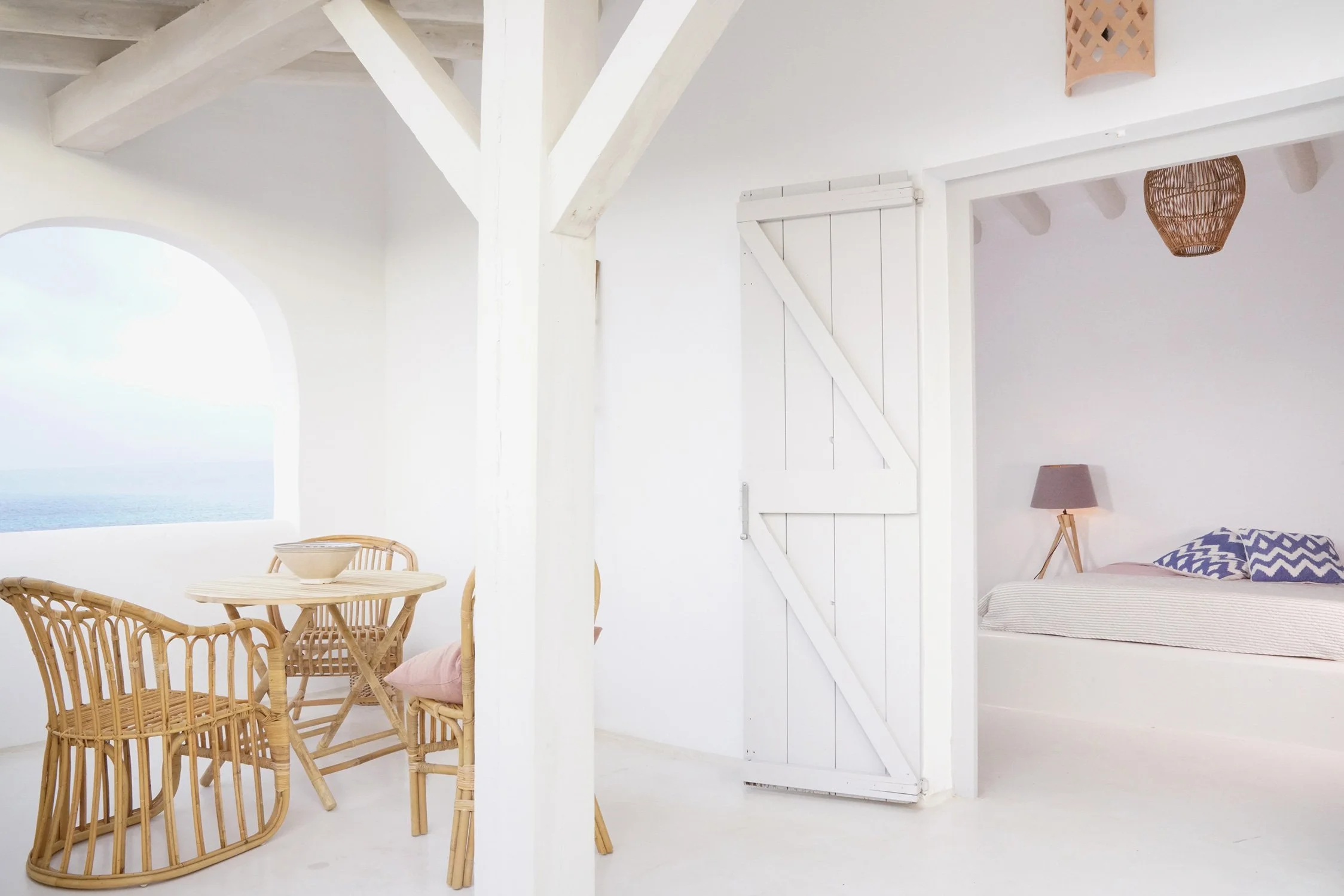 Intérieur minimaliste blanc avec coin repas en rotin, table ronde et chaises, et chambre à coucher avec lit, lampe de chevet et lampadaire en osier, vue partielle d'une fenêtre avec vue océan.