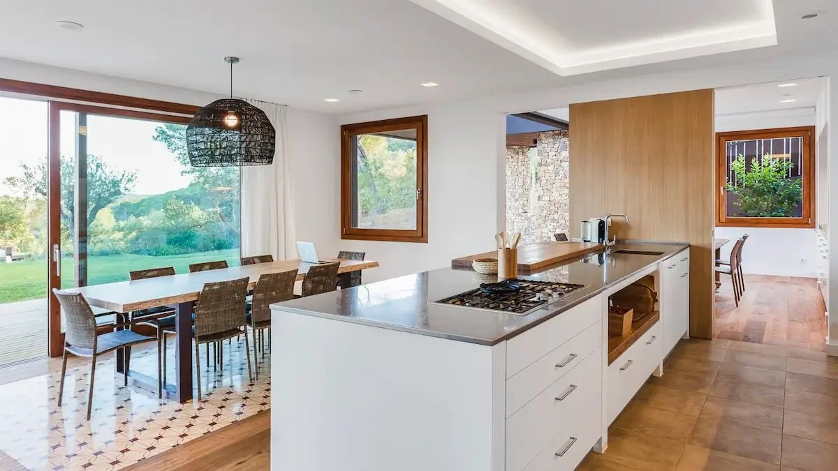 Intérieur lumineux d'une cuisine moderne avec coin repas, grande fenêtre, bois et blanc, terrasse extérieure visible.