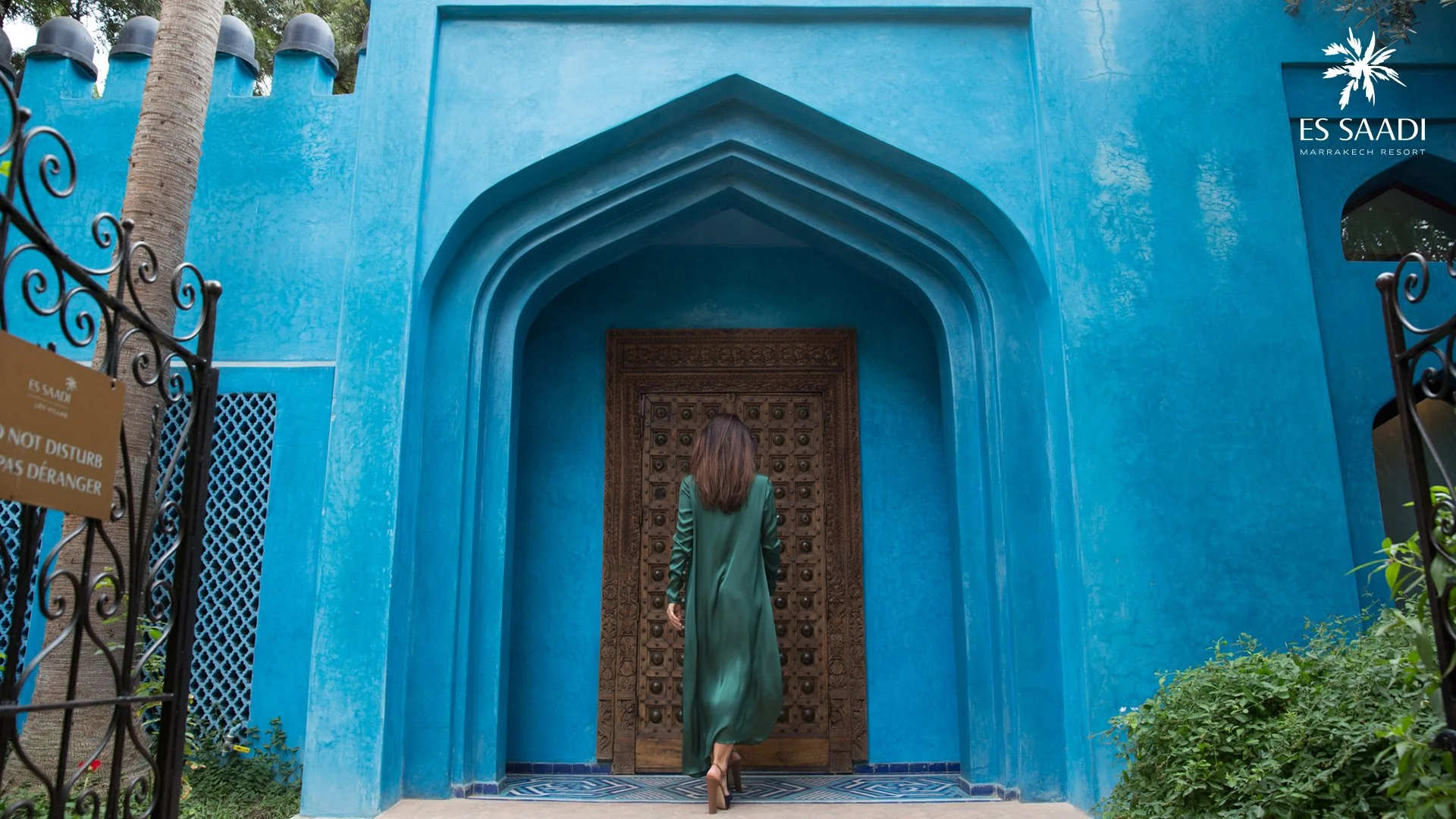 Une femme en robe verte entre dans un bâtiment marocain avec une porte en bois ornée, murs bleu vif, entouré de végétation, au rez-de-chaussée du complexe hôtelier ES SAADI à Marrakech.