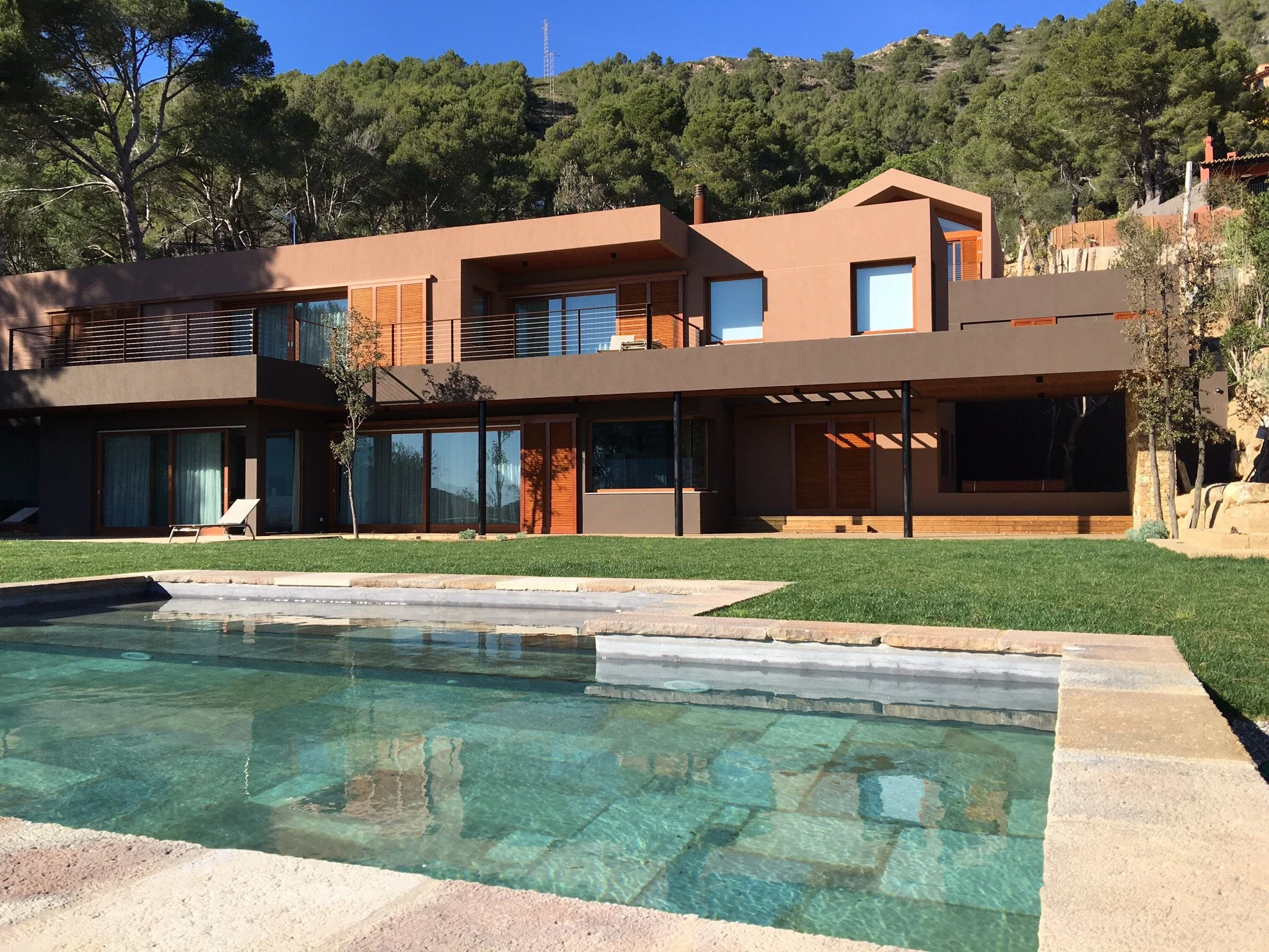 Maison moderne avec plusieurs étages, grande terrasse, piscine devant, dans un environnement naturel entouré d'arbres.