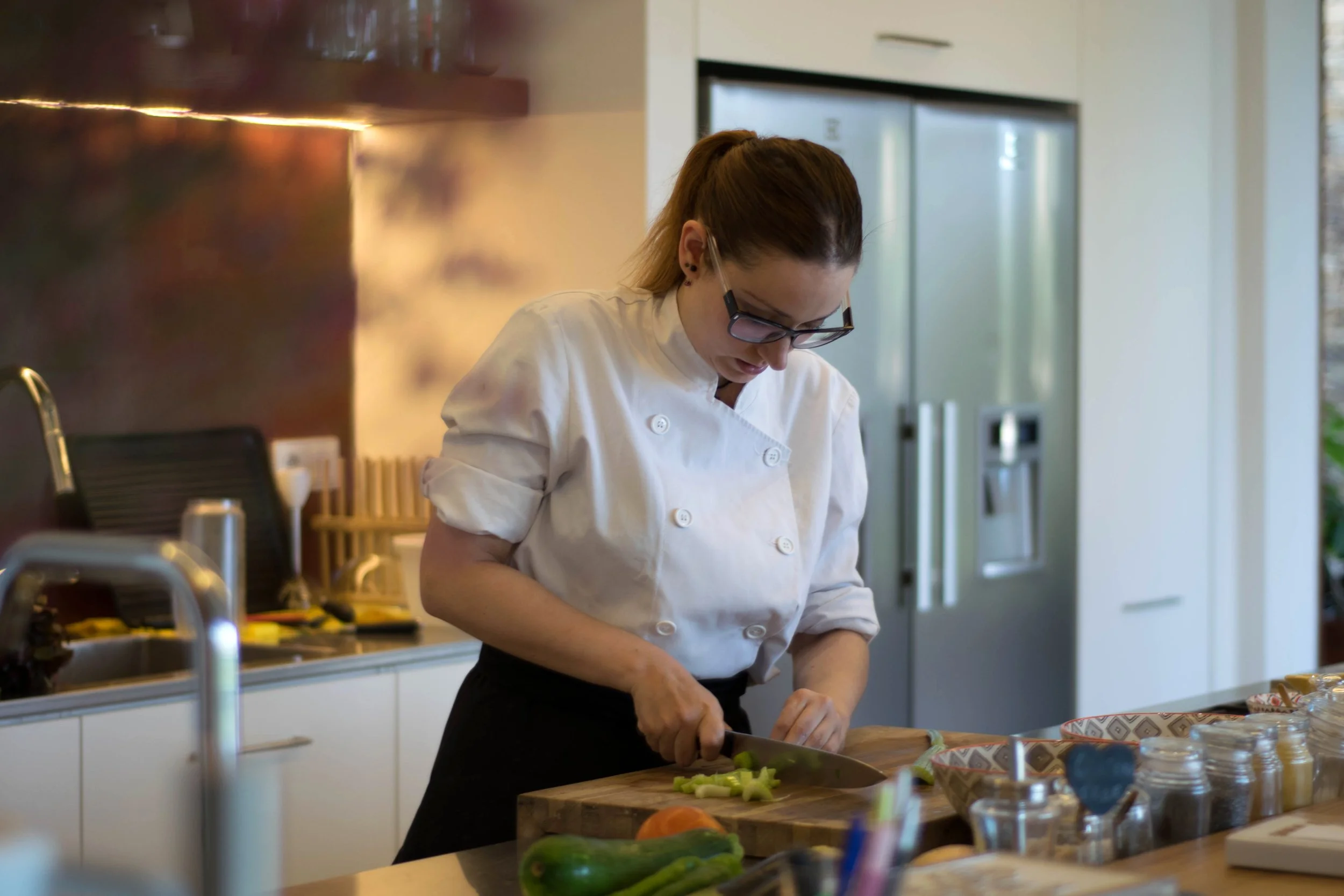 Une femme en uniforme de chef cuisinier coupe des légumes sur une planche à découper dans une cuisine moderne.