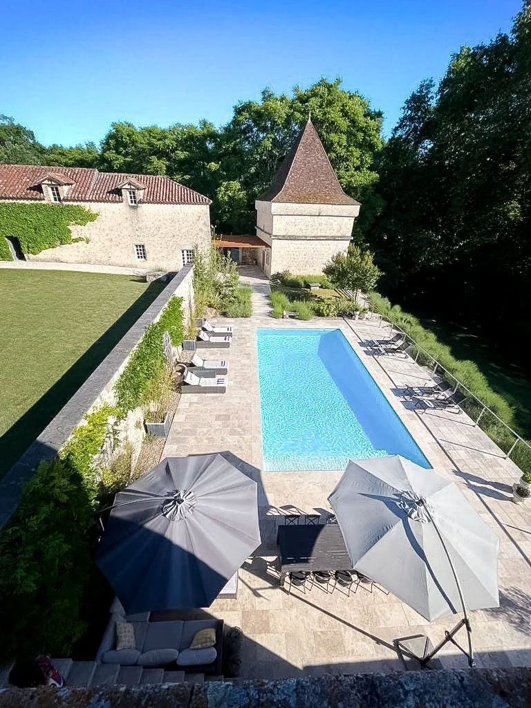 Piscine rectangulaire dans un jardin avec des parasols, chaises longues, un salon en plein air, un mur végétal, un vieux bâtiment avec une tour en pierre, et beaucoup d'arbres autour.