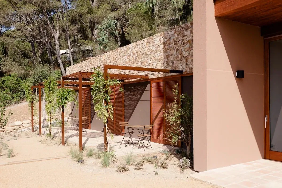 Terrasse extérieure avec pergola en bois, tables et chaises en métal, entourée de végétation, avec une maison moderne en arrière-plan dans un environnement naturel.