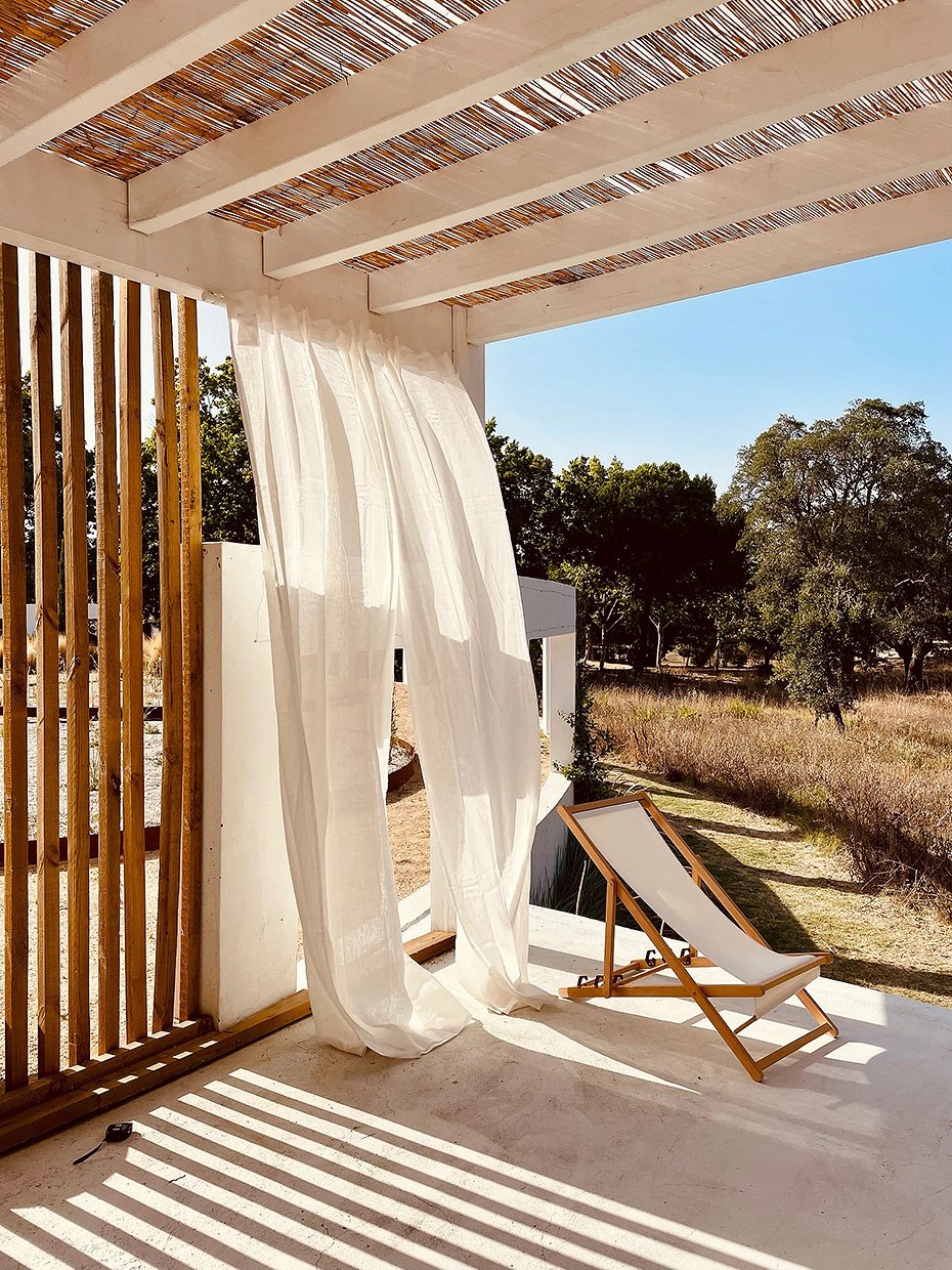 Une terrasse en plein air avec une chaise longue en bois et toile blanche, des rideaux légers blancs, un plafond en bois avec des bâtons de bois, et un paysage de campagne avec des arbres et un ciel bleu.