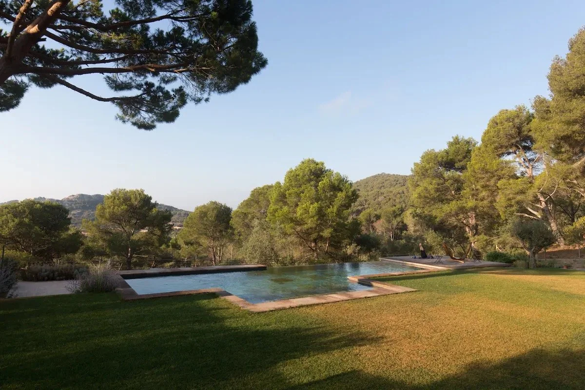 Piscine rectangulaire dans un jardin avec des arbres et des collines en arrière-plan, ciel clair.