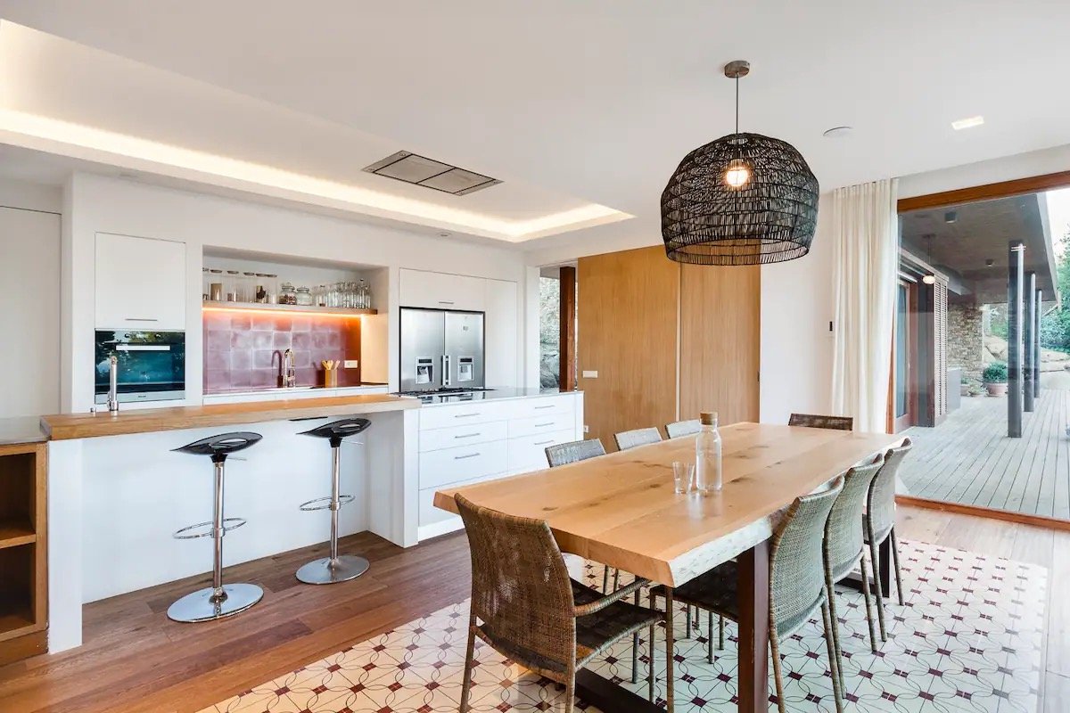 Intérieur d'une salle à manger et cuisine moderne avec une table en bois, chaises en rotin, un plafonnier en osier, un sol en carrelage et une ouverture donnant sur une terrasse en bois.