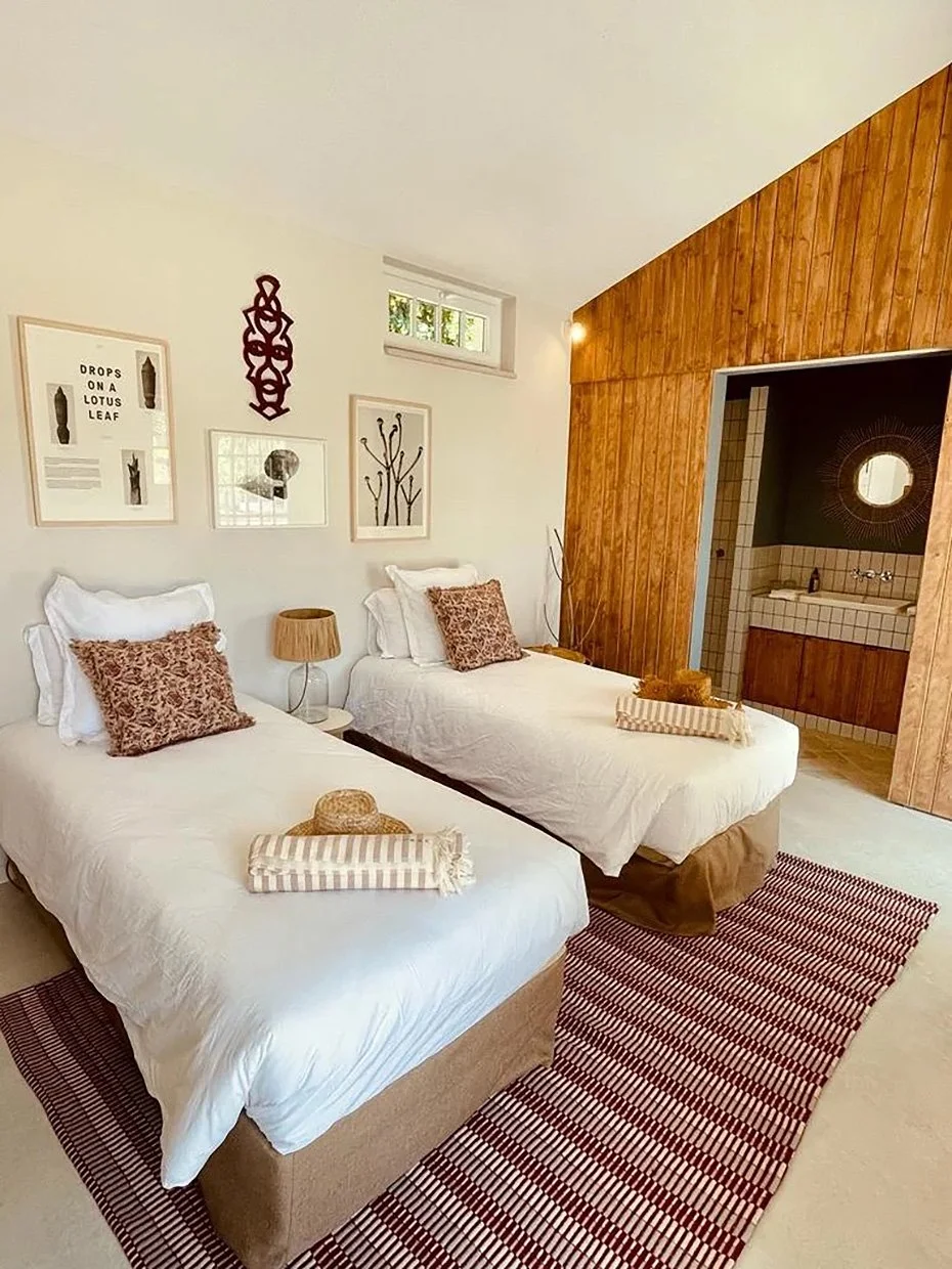 Chambre avec deux lits doubles, décoration moderne avec tableaux, tapis rayé rouge et blanc, petite salle de bain en bois visible à travers une ouverture.