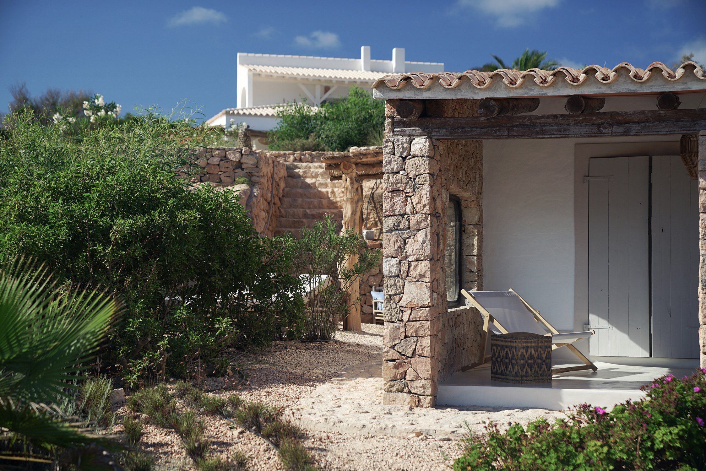 Terrasse en plein air d'une maison en pierre avec une chaise longue et un petit tabouret, entourée de végétation et sous un ciel bleu.
