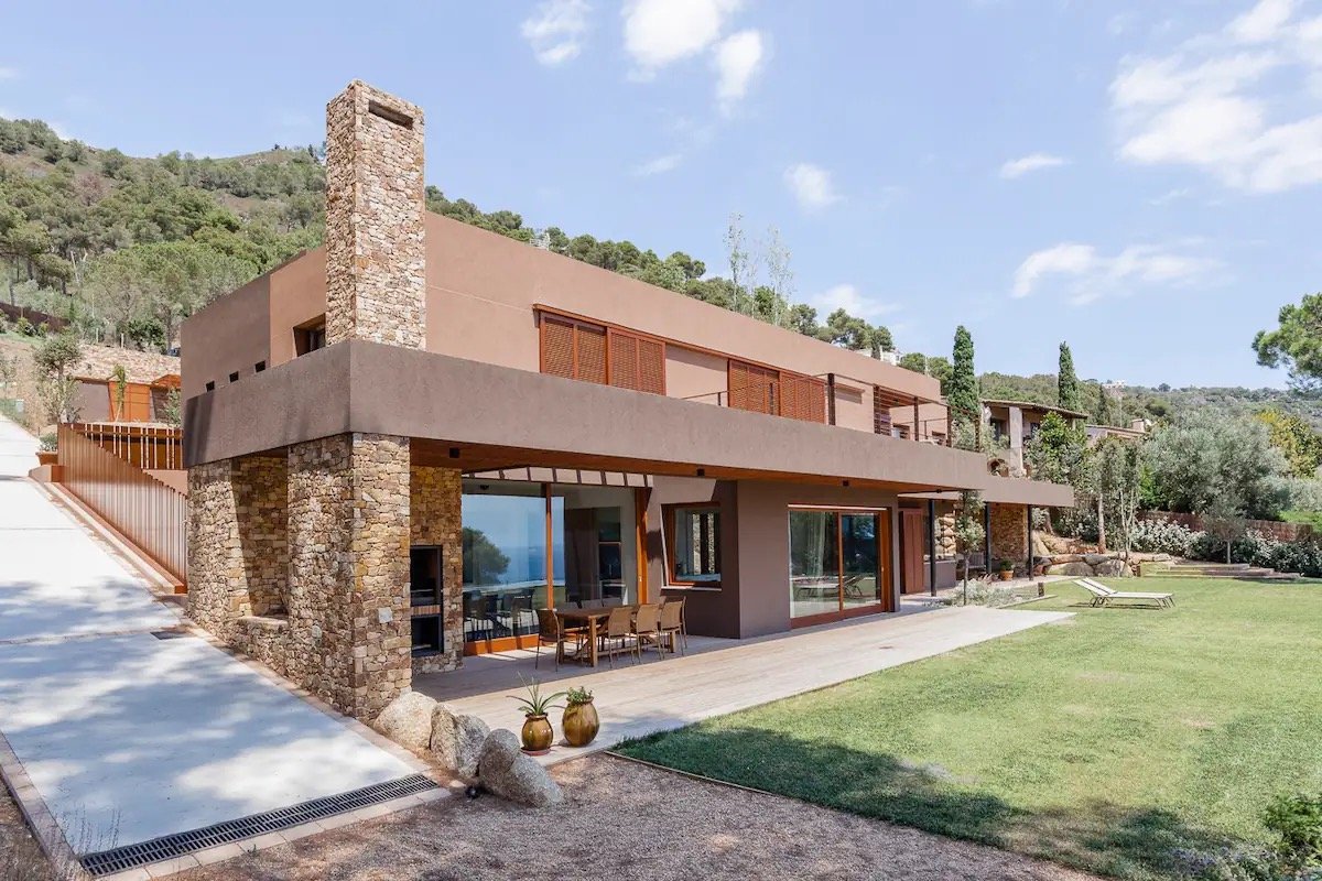 Une maison moderne avec un mélange de pierre et de matériaux lisses, grande terrasse avec mobilier en bois, entourée de végétation et de collines sous un ciel bleu.