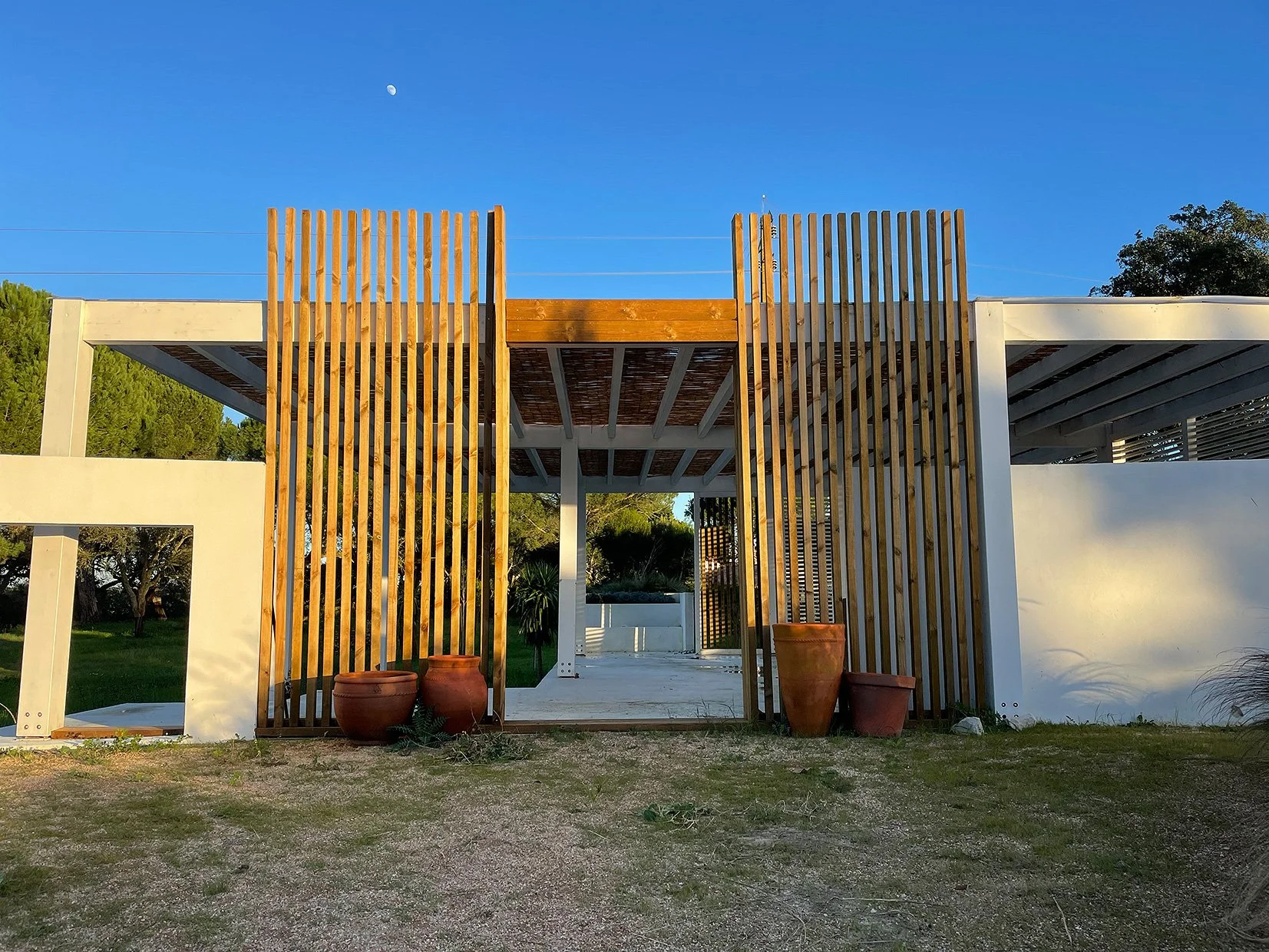 Une maison moderne avec une structure en bois et en béton, un ciel bleu clair, la lune visible, et plusieurs pots en terre cuite devant l'entrée.