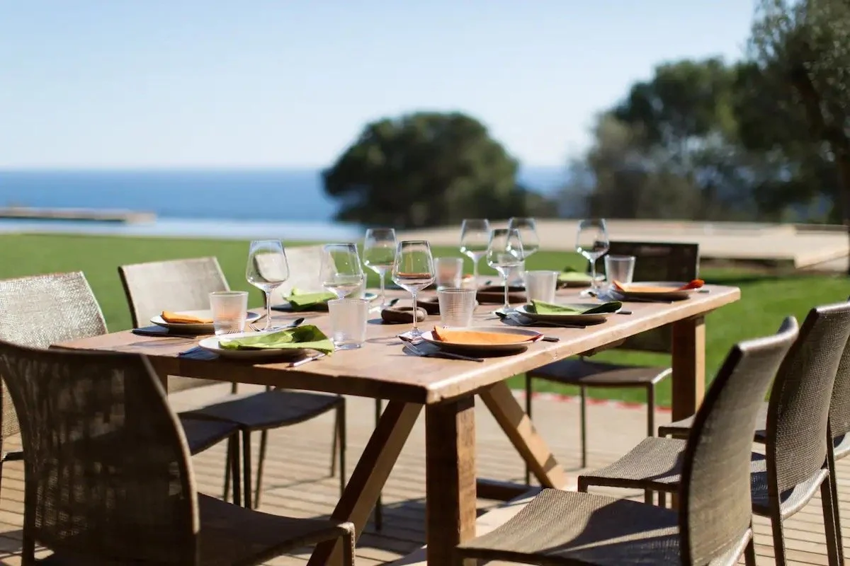 Table de restaurant en extérieur avec plusieurs verres à vin, assiettes et couverts, sous un ciel dégagé avec vue sur la mer et des arbres en arrière-plan.