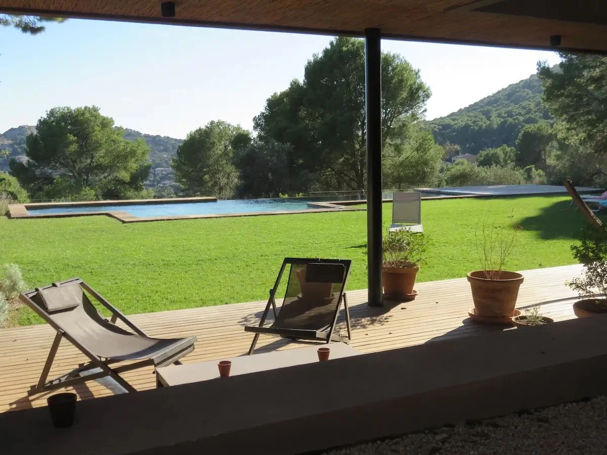 Terrasse avec chaise longue, plants en pot, vue d'une piscine et de collines verdoyantes en arrière-plan.