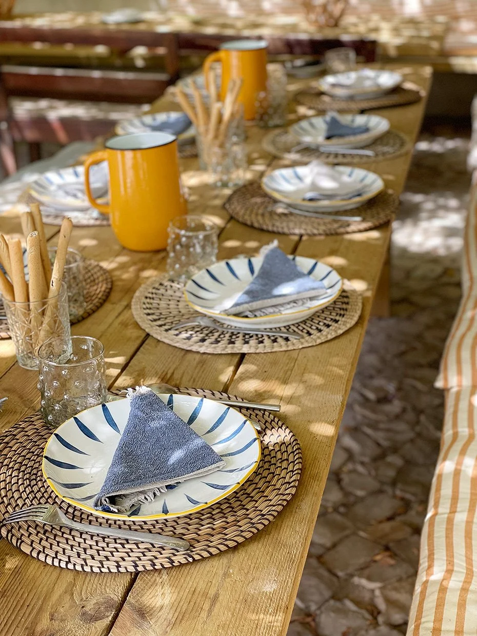 Une table en bois décorée pour un repas en plein air avec des assiettes, des verres, des couverts, des serviettes pliées, des gobelets en verre et deux mugs jaunes.