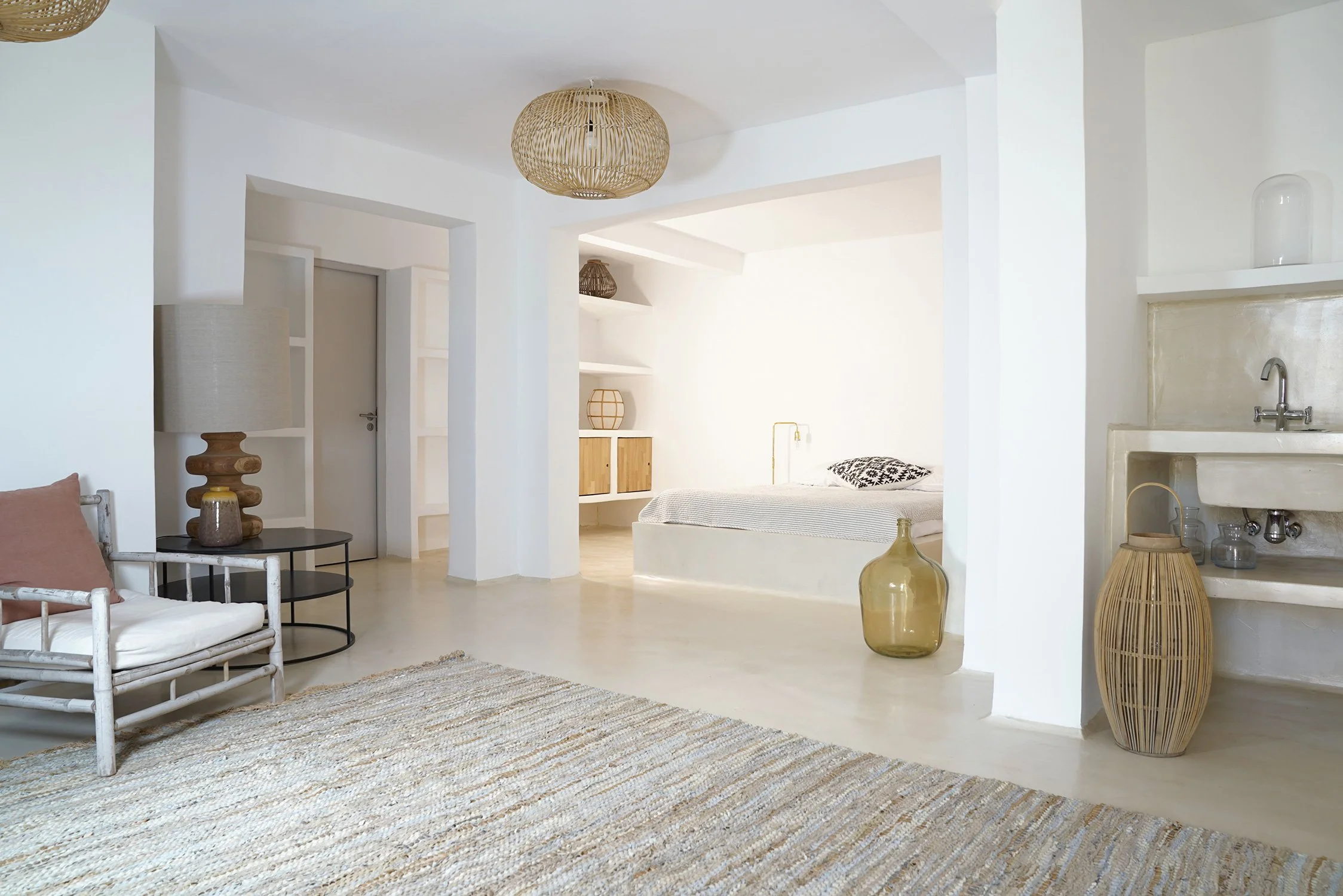 Un intérieur de maison moderne avec des murs blancs, un tapis beige, un canapé blanc avec un coussin rose, une lampe en bois sur une table ronde, un grand vase beige, une chambre avec un lit blanc, des étagères et une vasque.