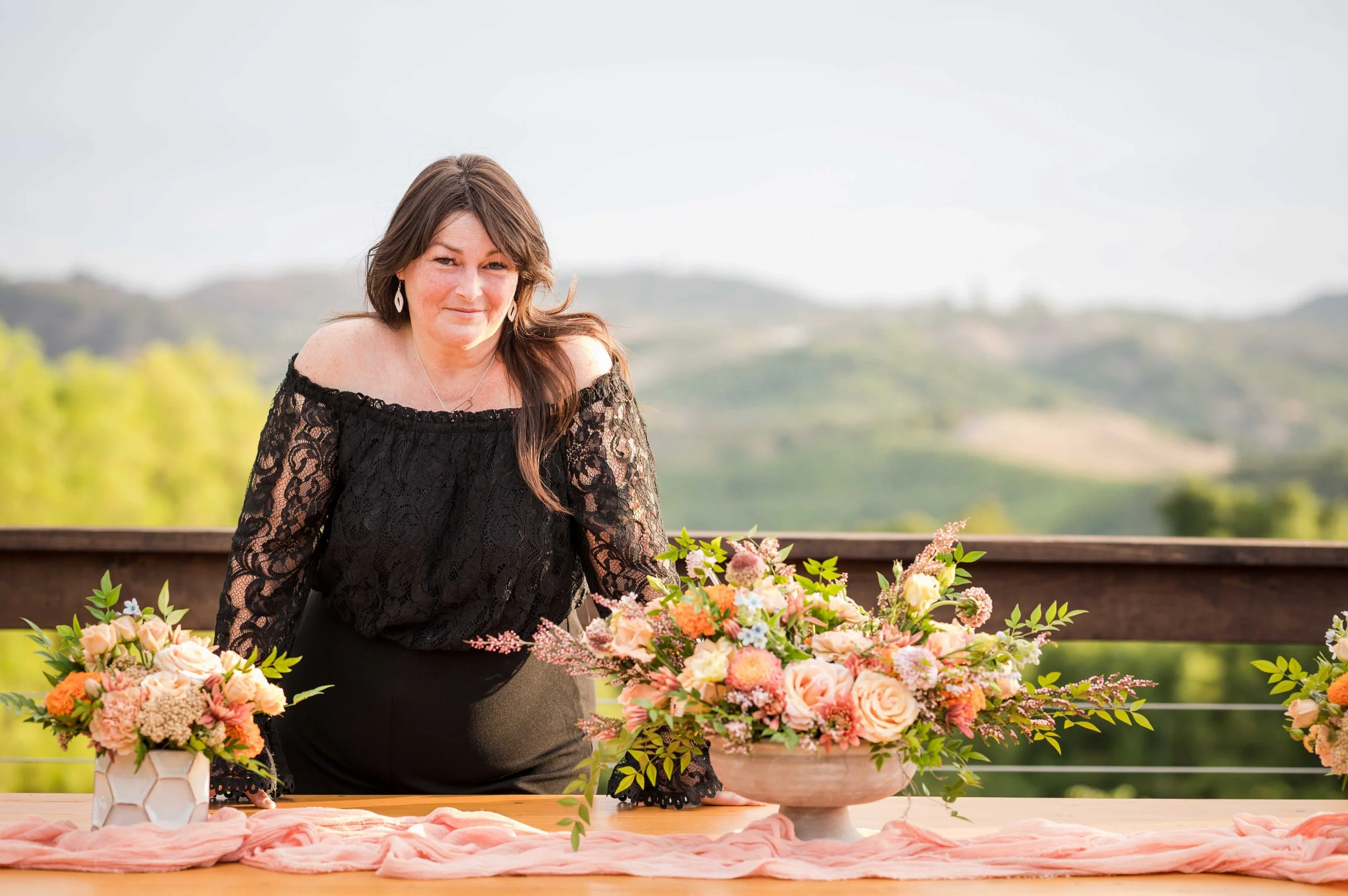 Coordinator and Floral Designer Rainy Diane standing behind a summer flower table arrangement. Peach and orange local grown flowers in a winnie compote centerpiece. The green rolling hills of Fallbrook fade away against the afternoon sky.