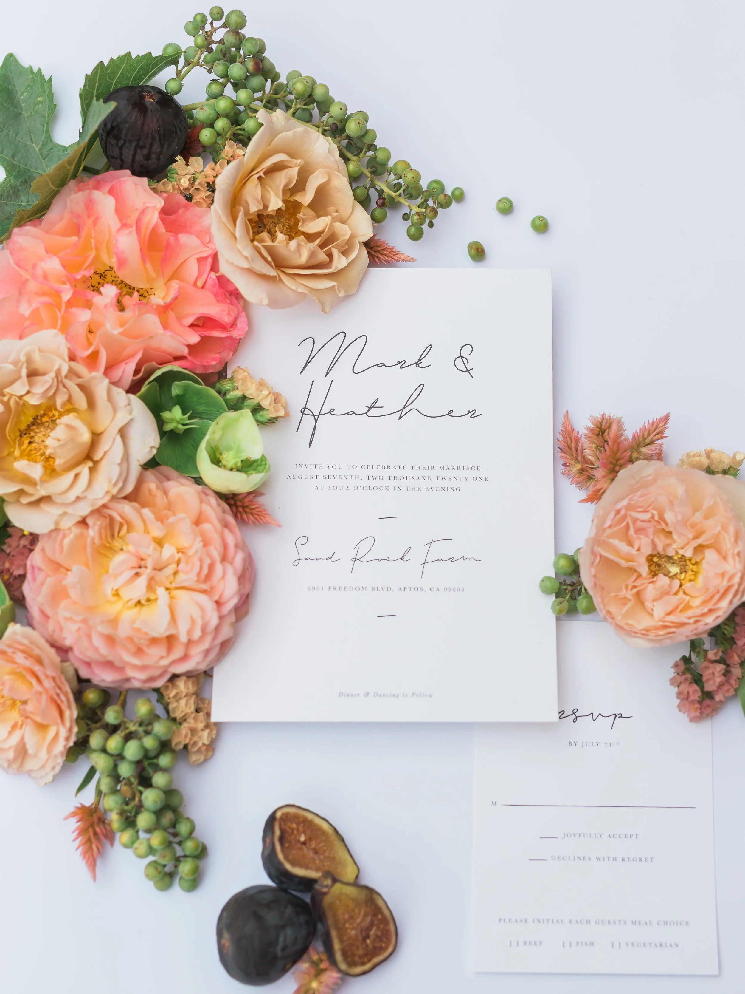 Wedding invitation surrounded by peach and pink flowers, green foliage, a cluster of green berries, and figs.