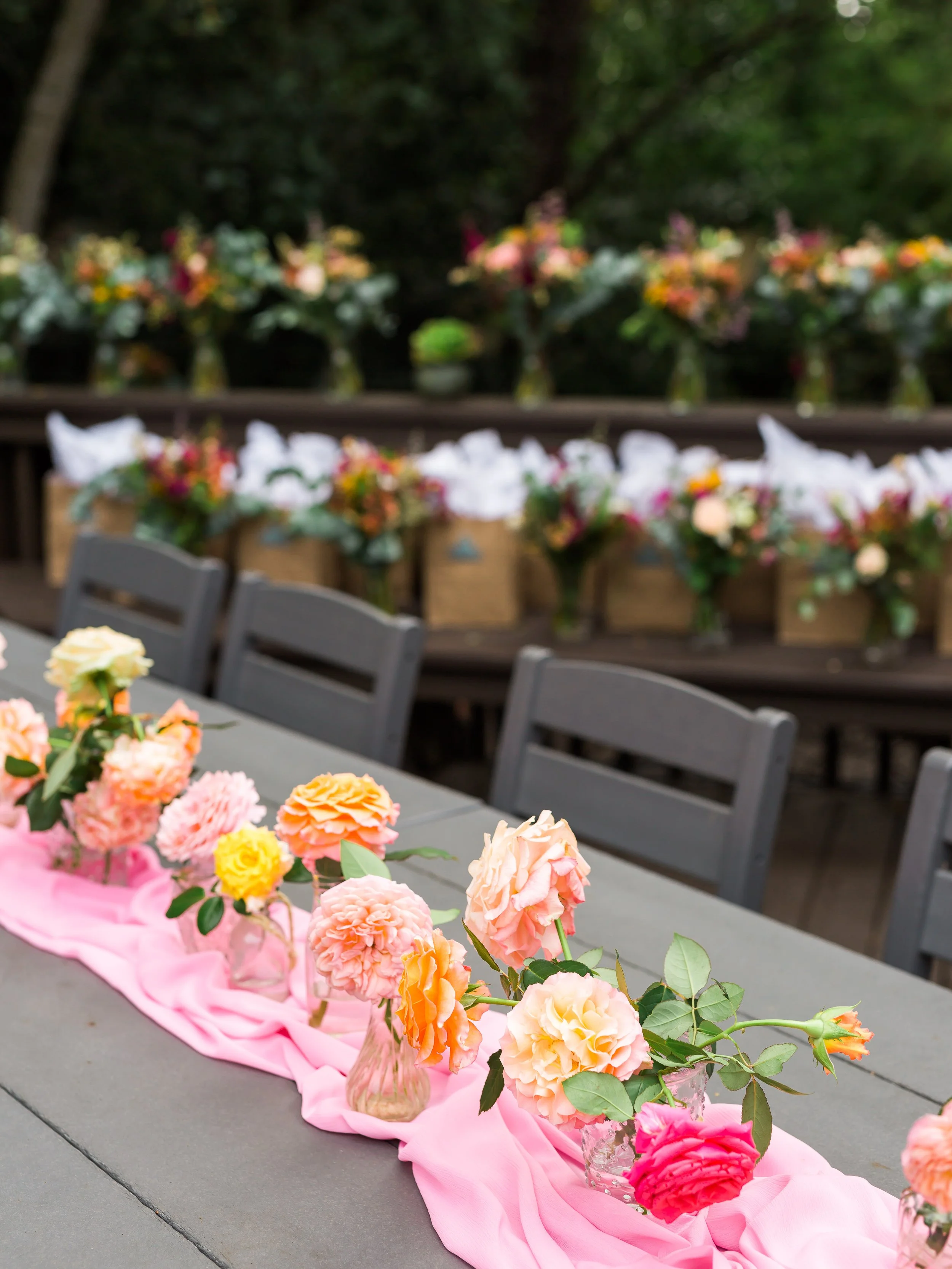 Arrangement of pink and peach roses in glass vases on a gray table with pink cloth, outdoor setting with additional flowers in the background.