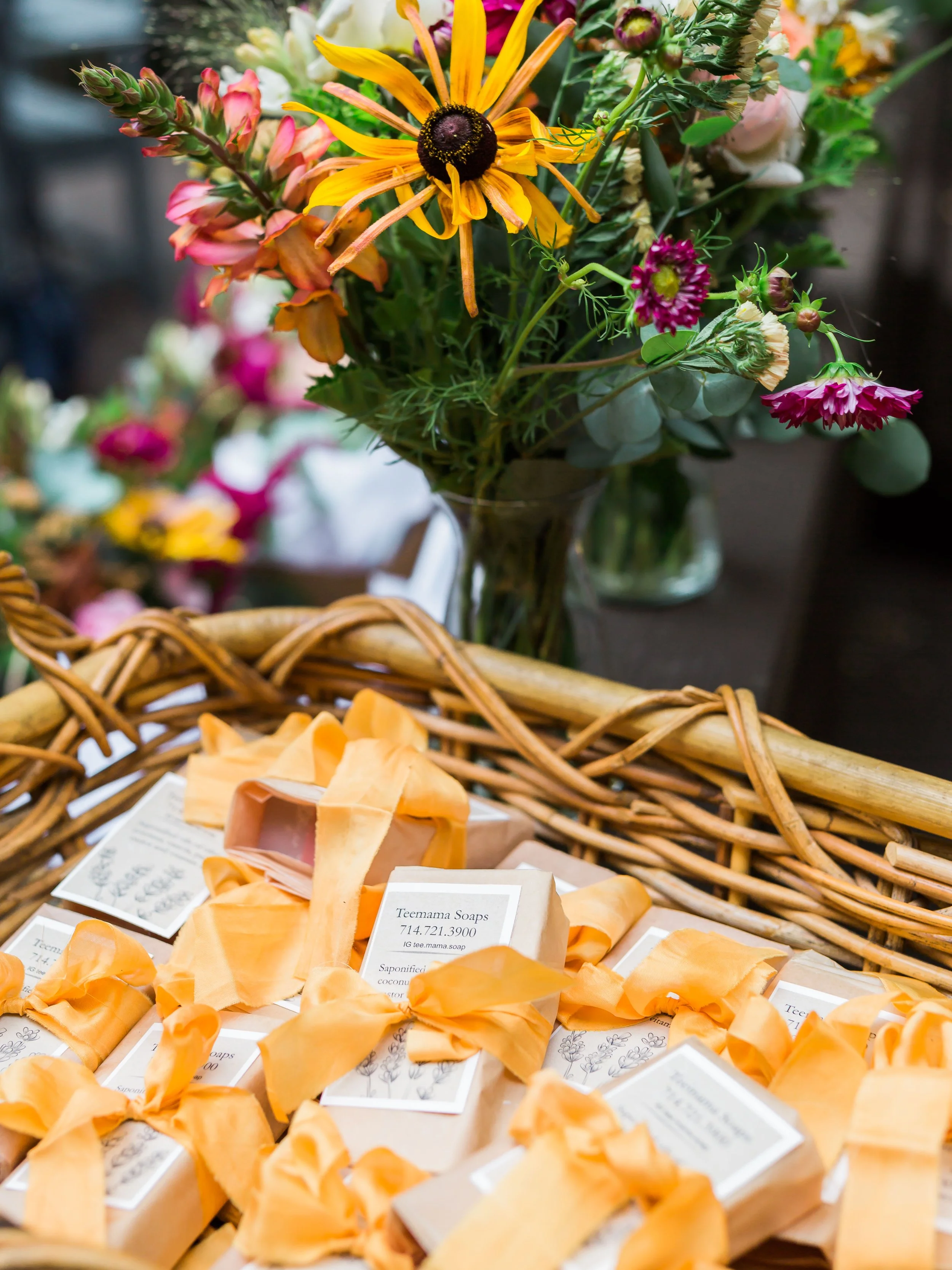 A basket filled with soap bars wrapped in orange paper with yellow ribbons sits in front of a vase of colorful flowers, including yellow, pink, and purple blossoms.