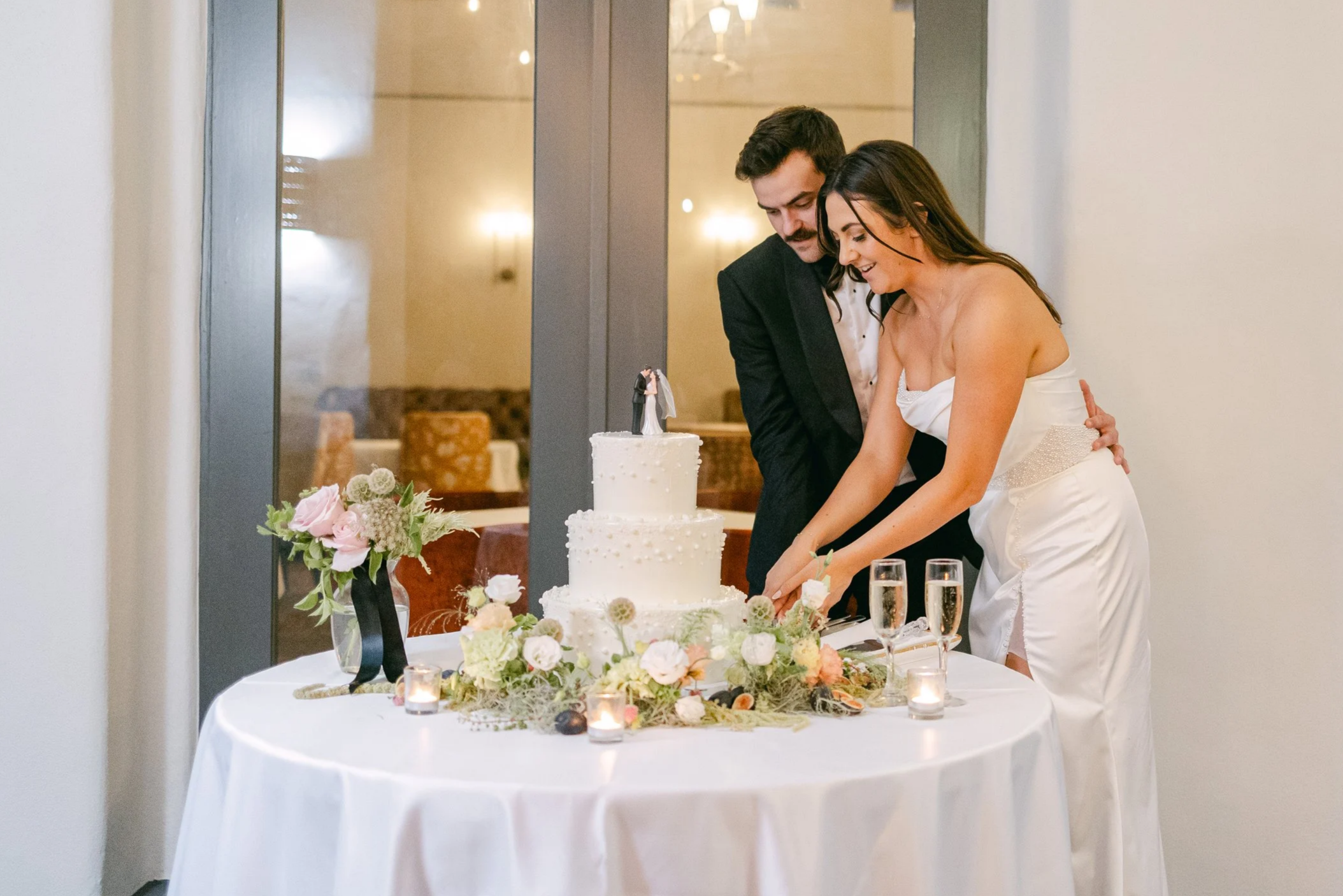 the happy couple cuts thier wedding cake. Simple garden meadow in light greens and blush florals, pops of dark figs  accents the table.