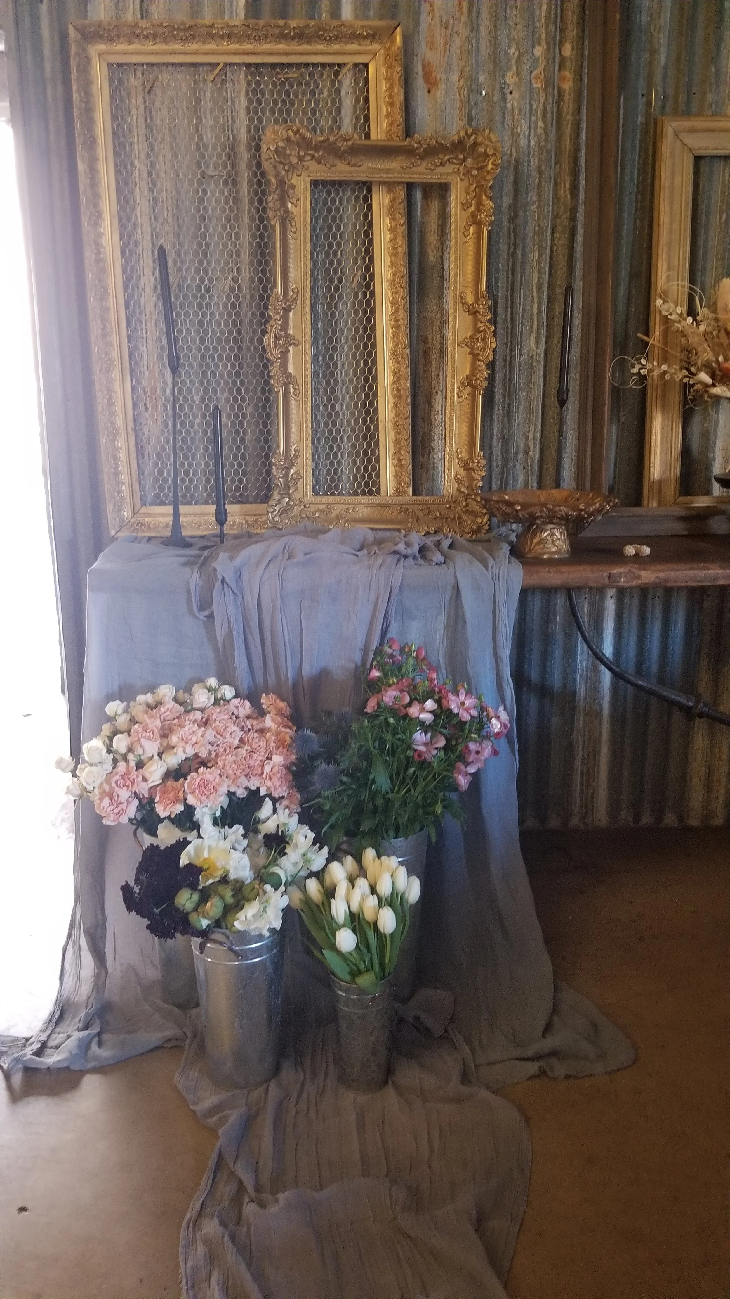 Decorative arrangement with cut flowers in metal vases, an ornate gold picture frame, and an empty rectangular gold frame, set against a rustic wood wall with a cloth covering a table or surface.