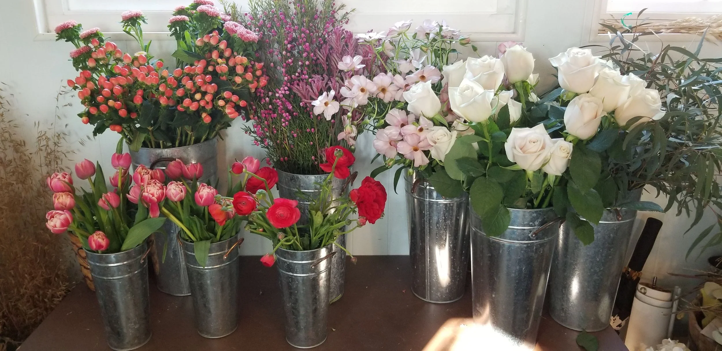 Assorted fresh flowers in metal buckets on a table, including pink tulips, red ranunculus, white roses, pink daisies, and purple waxflowers.