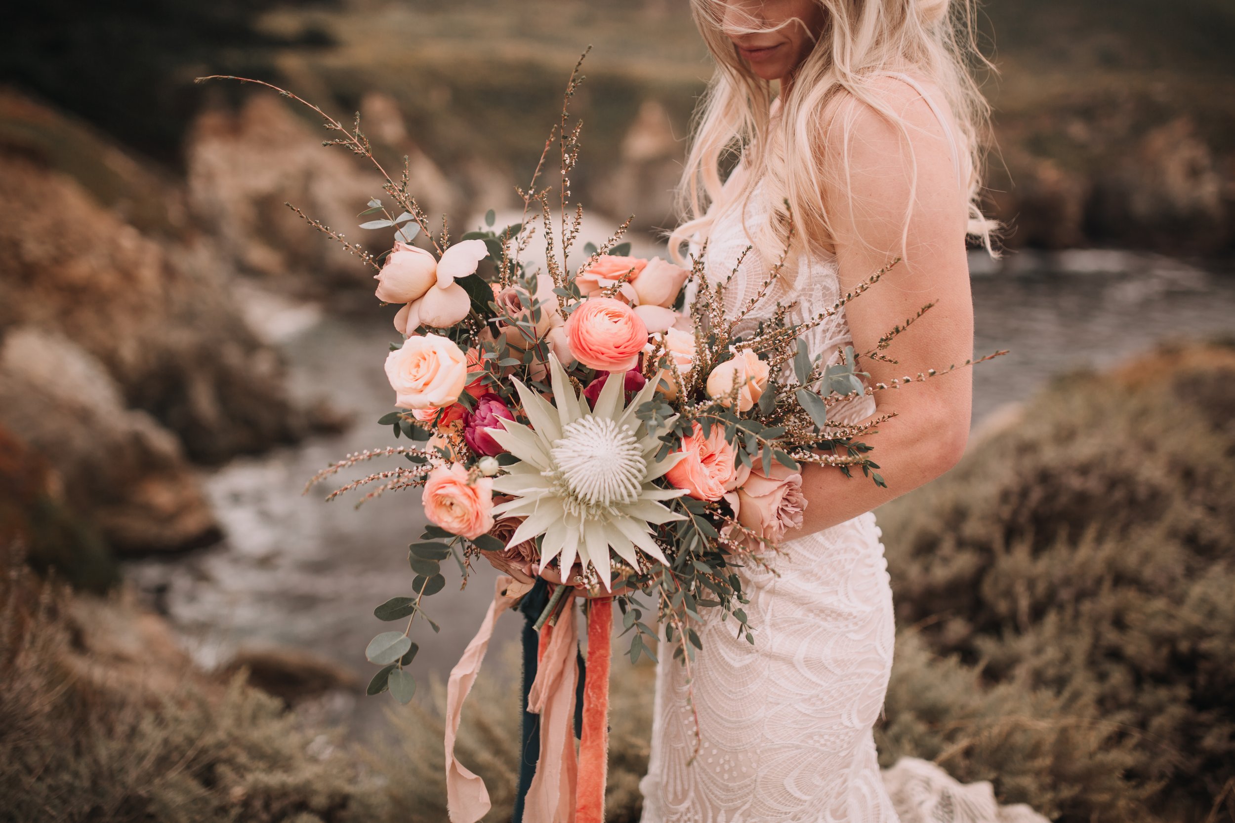 Big sur elopement  summer flower bouquet king protea ranunculus  garden roses. california coast background bride with flowy blonde hair