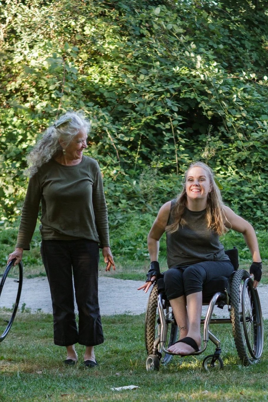 Two dancers together on grass with green trees in the background. The older woman has long gray hair and walks beside the younger woman with blonde hair  in a wheelchair. Both are wearing green tops, smiling and moving toward the camera.