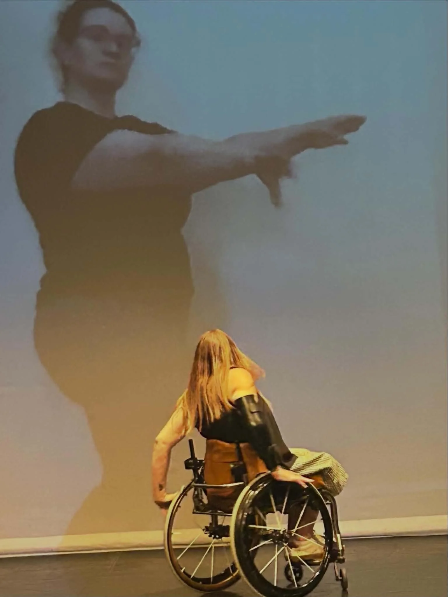 Harmanie, a wheelchair dancer, is in mid-turn, her hair fanning out in front of Kelsie, a standing dancer, projected three times Harmanie’s size on a screen.