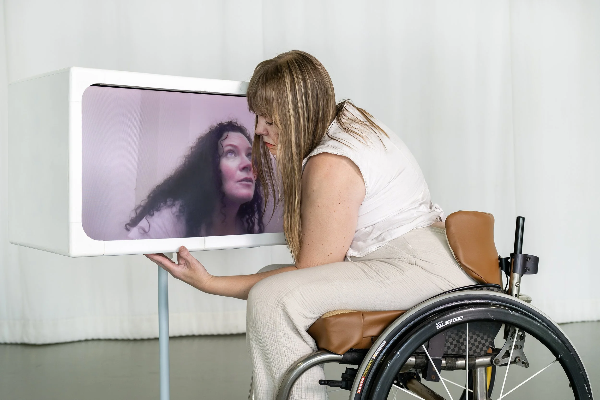 Harmanie a manual wheelchair users gently cradles a screen in a box called Echo. In the Box (in Echo) Kelsie with long curly hair looks up and out of the screen as if she can see into Harmanie's eyes.