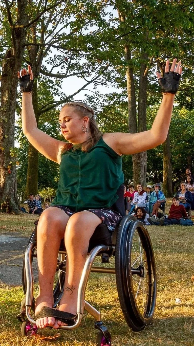 A dancer with long hair in a braid, wearing a green sleeveless top, floral skirt, and black gloves, is sitting in a wheelchair with her arms raised and eyes closed in a wooded park, with people sitting on the grass in the background.