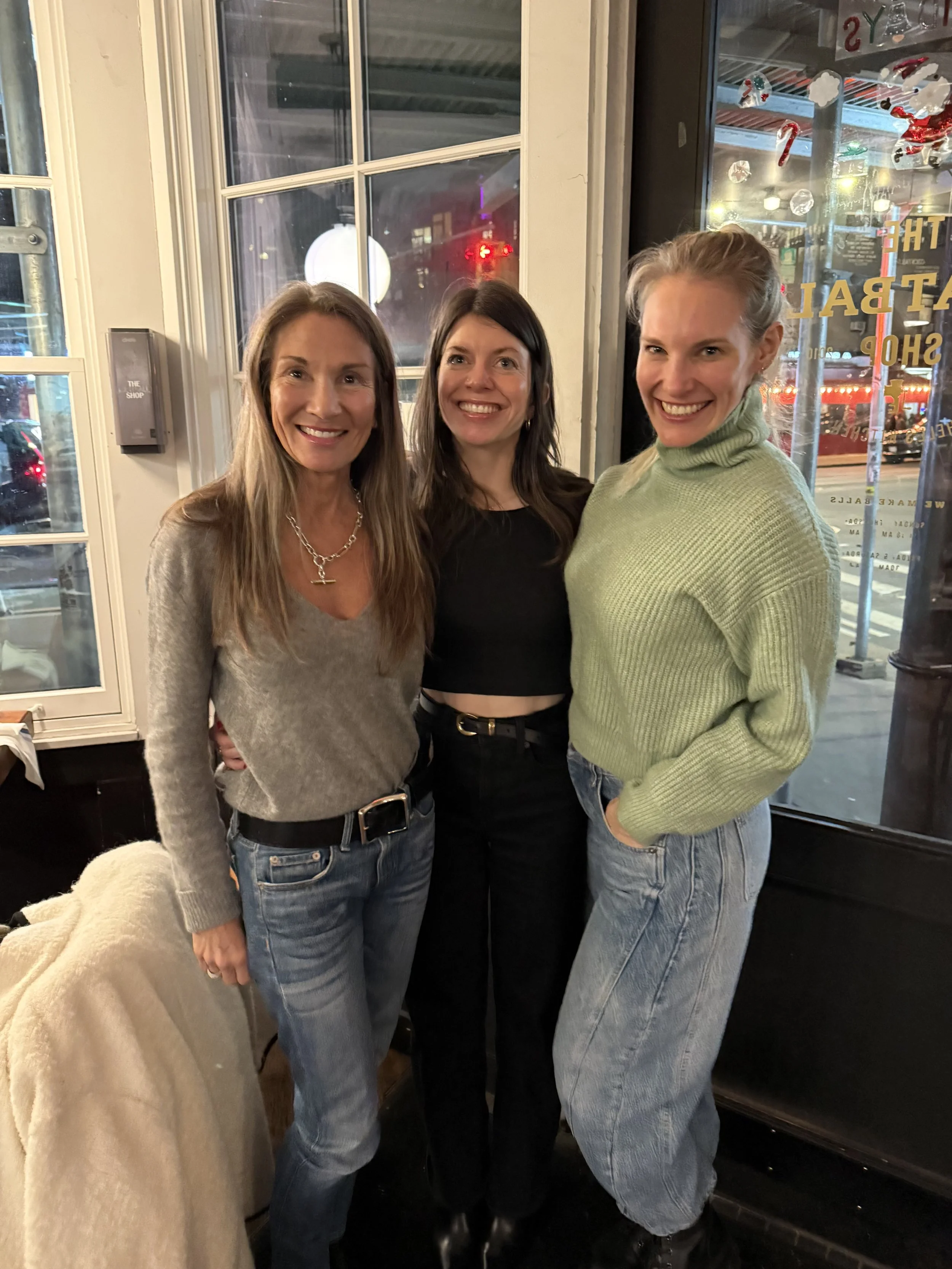 Three women smiling and posing together inside a restaurant or cafe near a window with city lights outside.