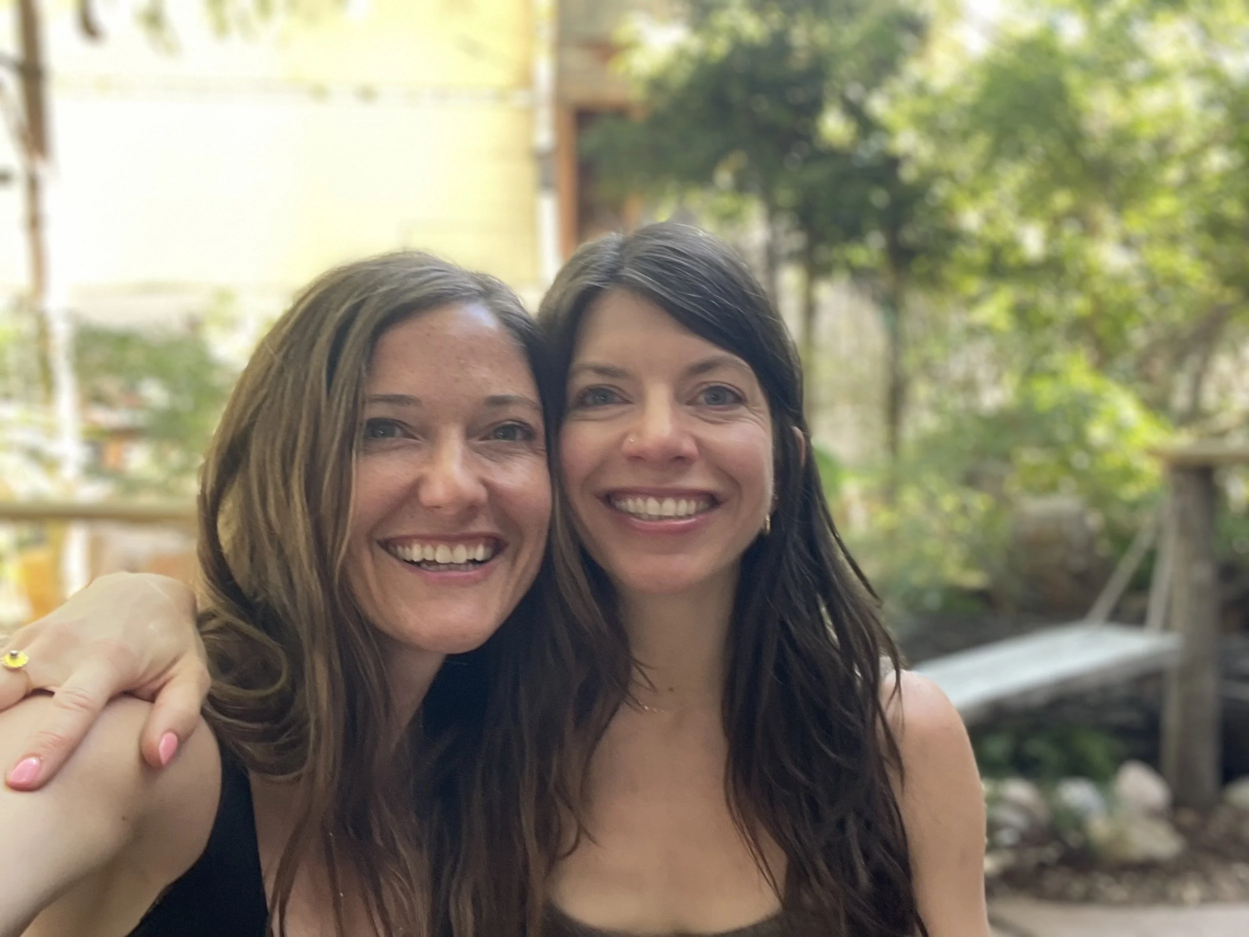 Two women smiling and posing together outdoors with a blurred background of trees and a fence.