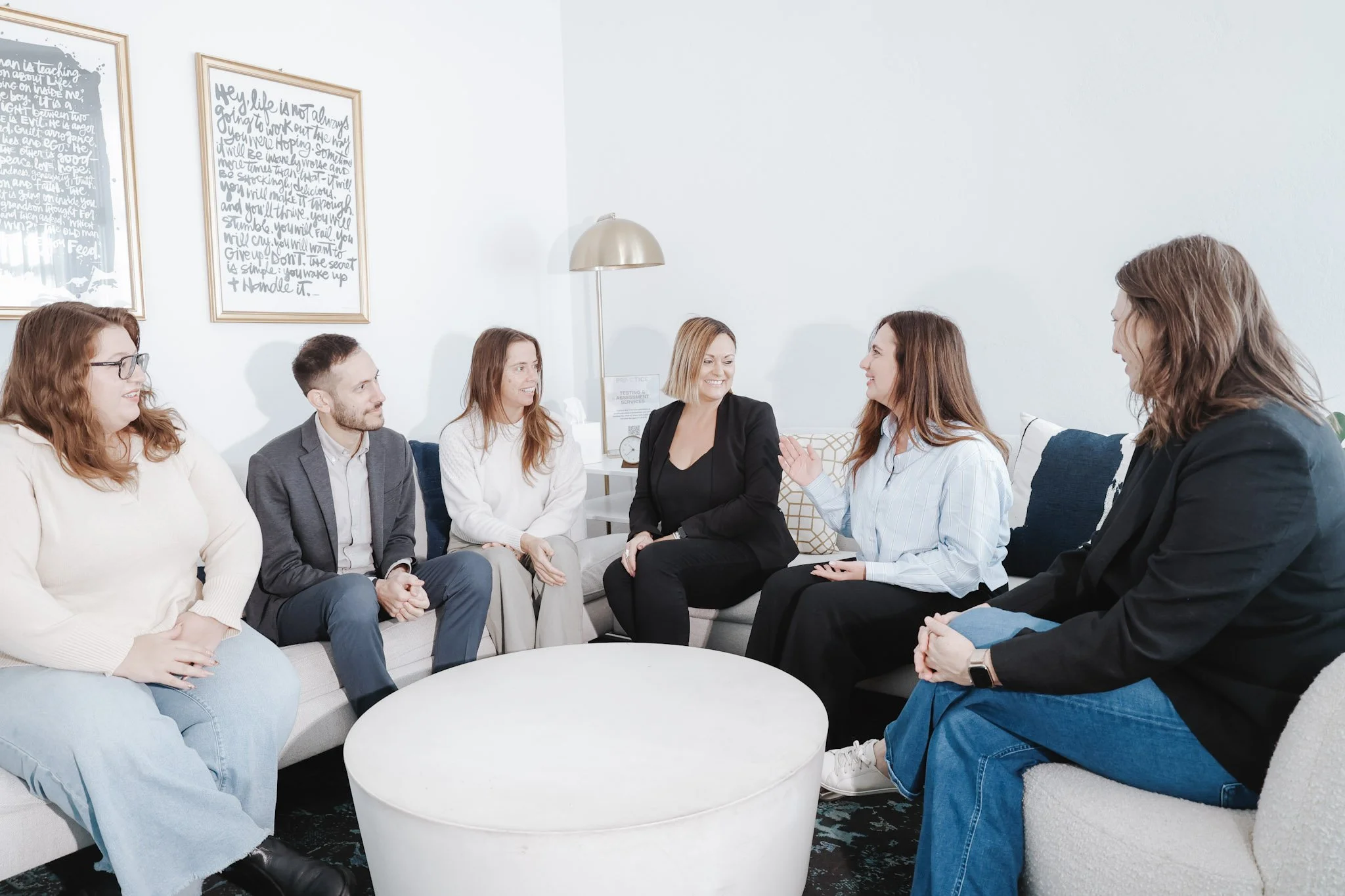A group of seven diverse people sitting on a sofa and chairs in a bright room, engaged in conversation, with framed quotes on the wall behind them.